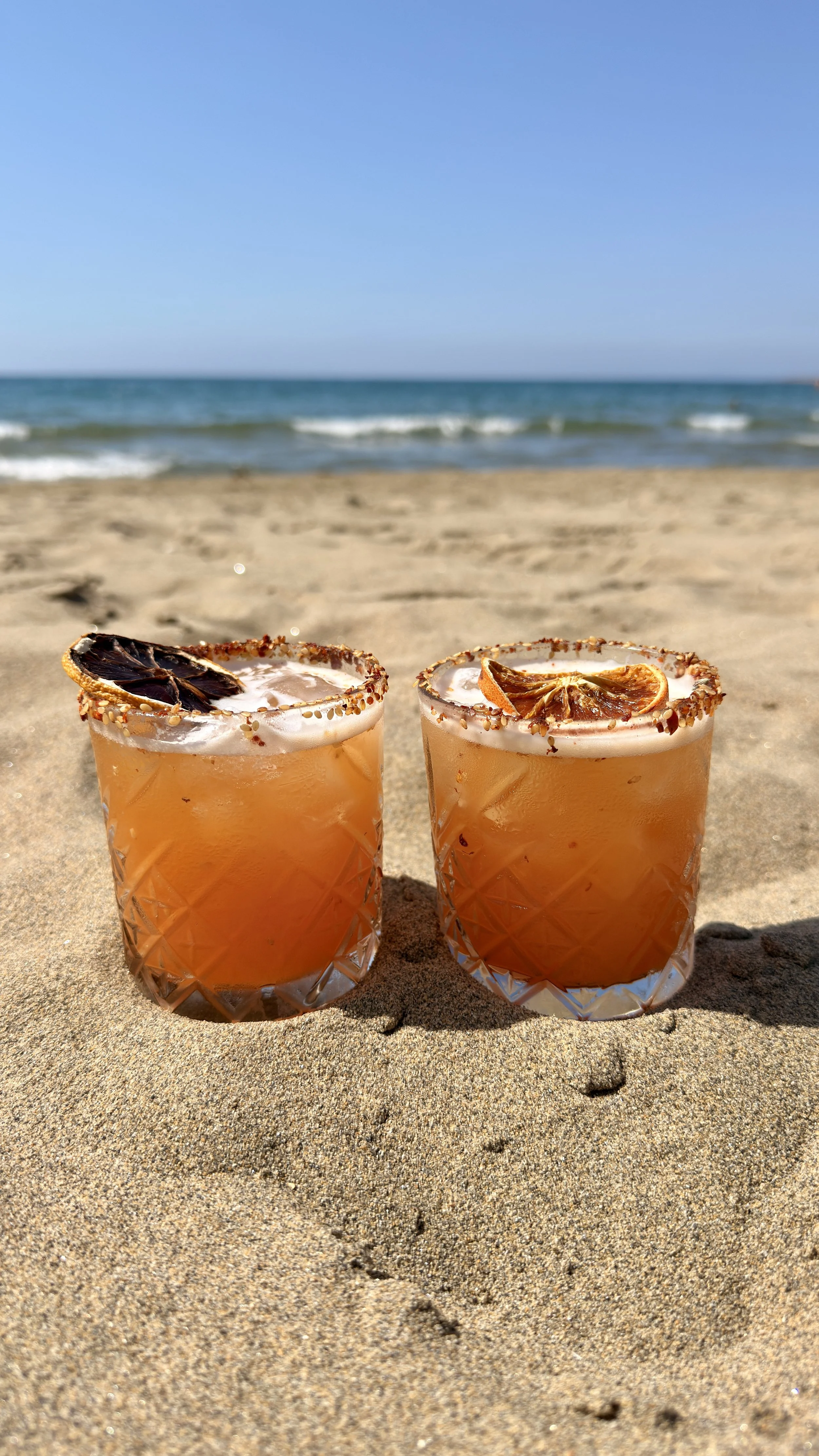 Two glasses of orange-colored cocktail with salted rim and dried citrus slices on top, placed on sandy beach with ocean waves and blue sky in the background.
