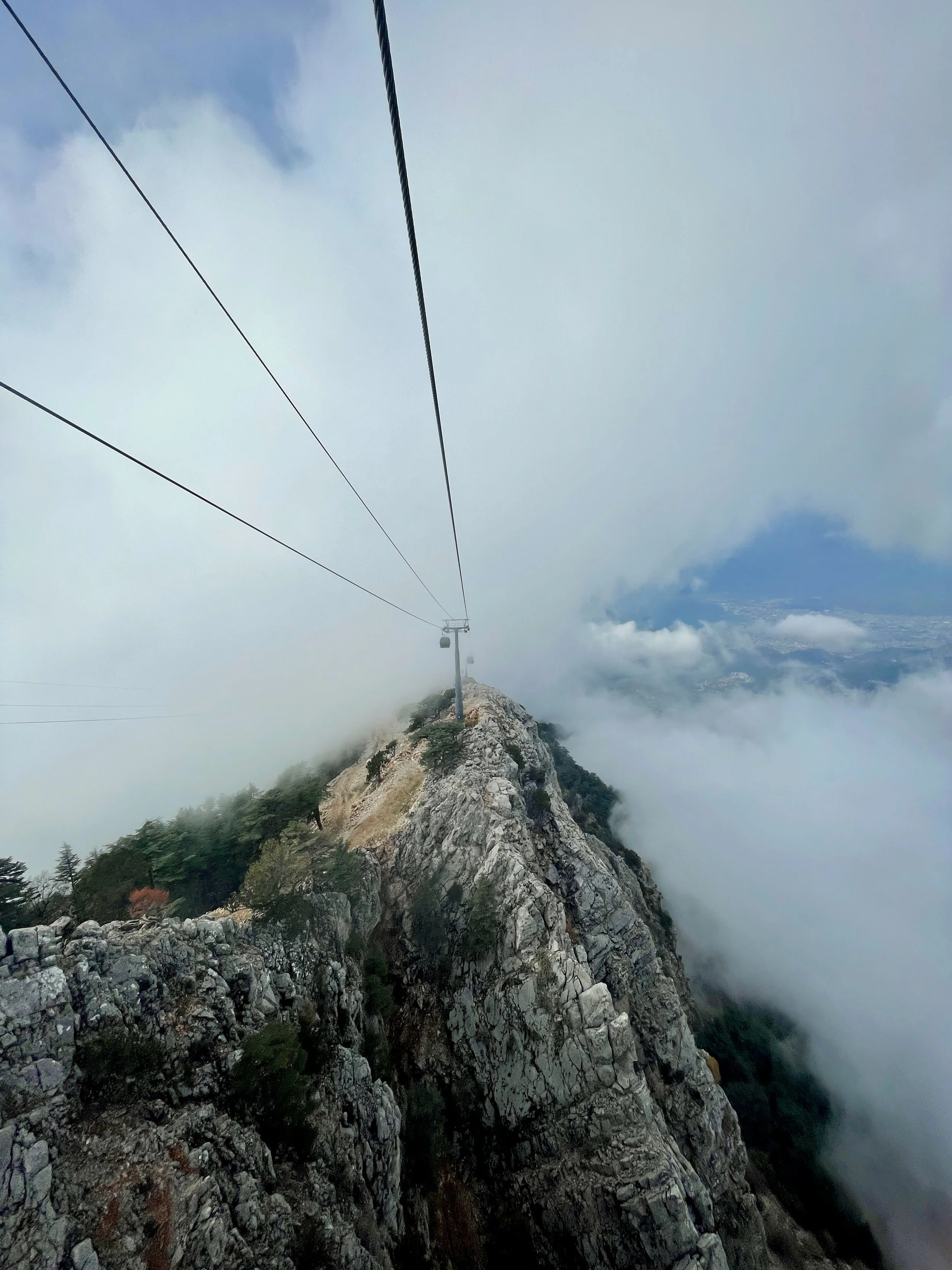 A mountain with rocky terrain and sparse trees, shrouded in clouds, with a cable car system extending over it into the foggy sky.