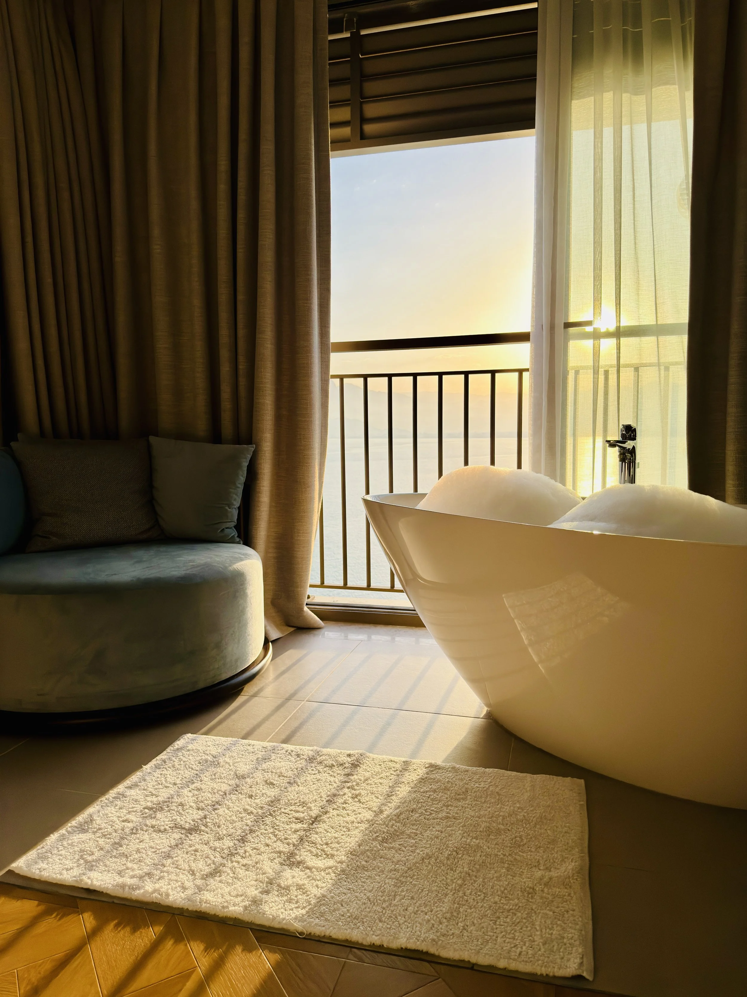 Sunlight coming through a balcony door with sheer curtains, a bathtub with a white towel, a beige sofa with pillows, and a white shag rug on a wooden floor.