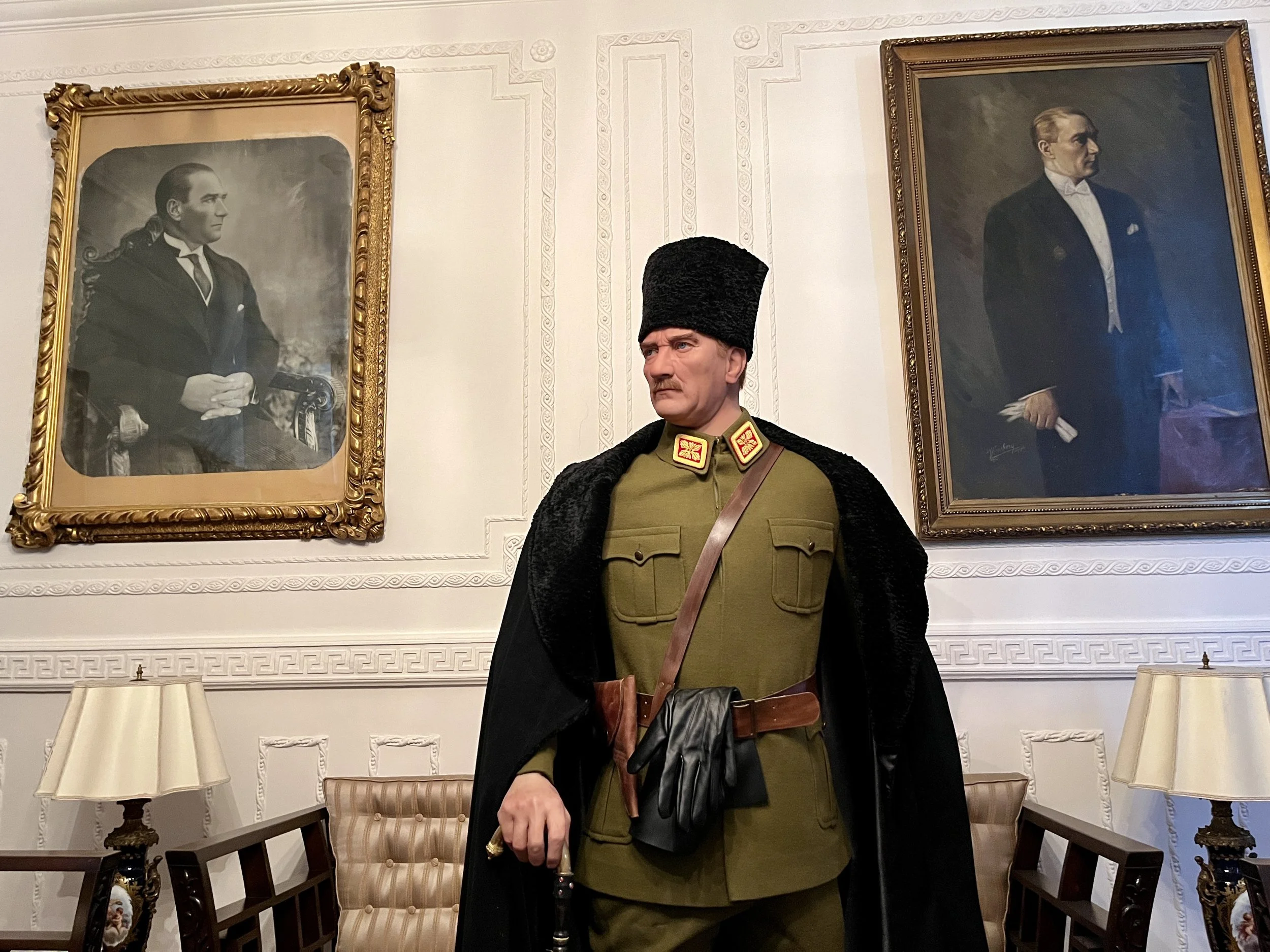 A wax figure of a military officer dressed in a green uniform with medals, black cape, and a tall black fur hat, standing in a room with framed portraits of historical figures on the wall.