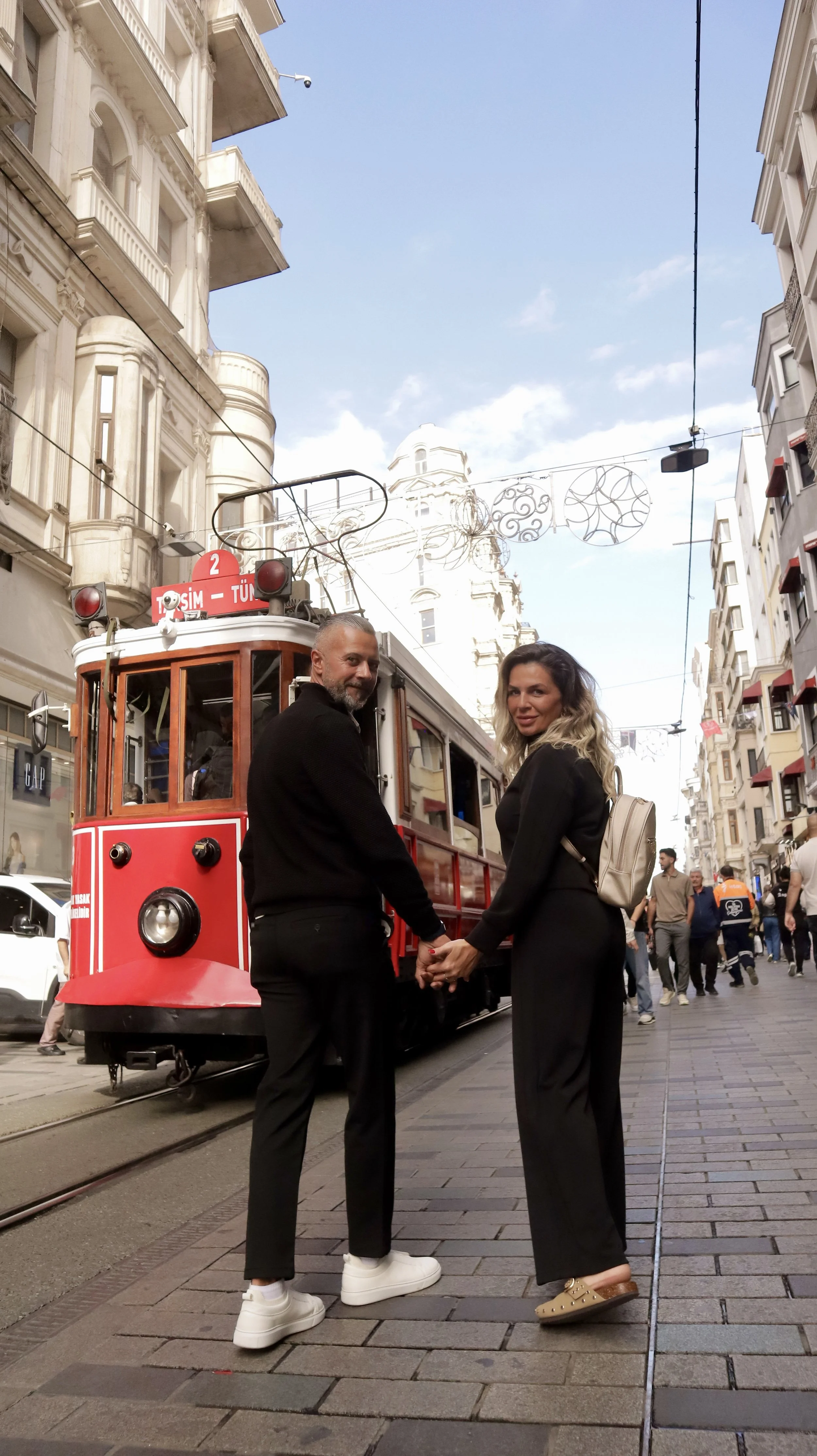 A couple holding hands and smiling at each other on a city street with a vintage red tram in the background. The street is busy with pedestrians and lined with buildings.