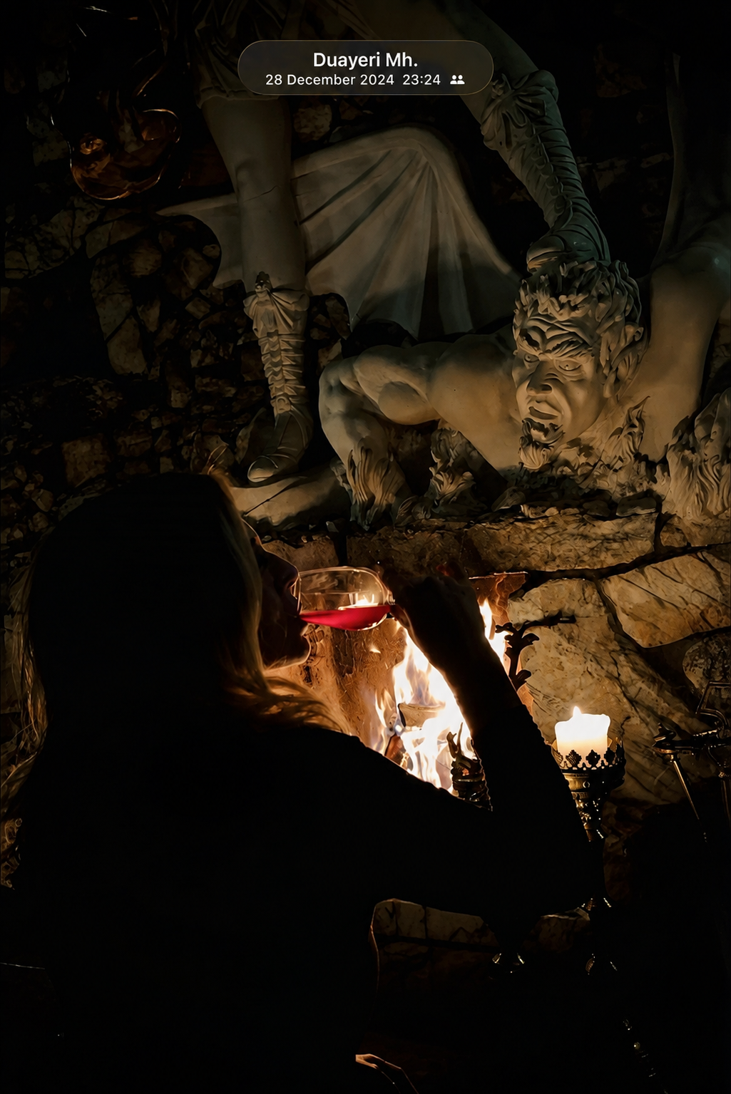 A woman with long hair drinking red wine from a glass, sitting by a fireplace with flames, with a large stone sculpture of a mythical creature with wings and a fierce face on the wall behind her.