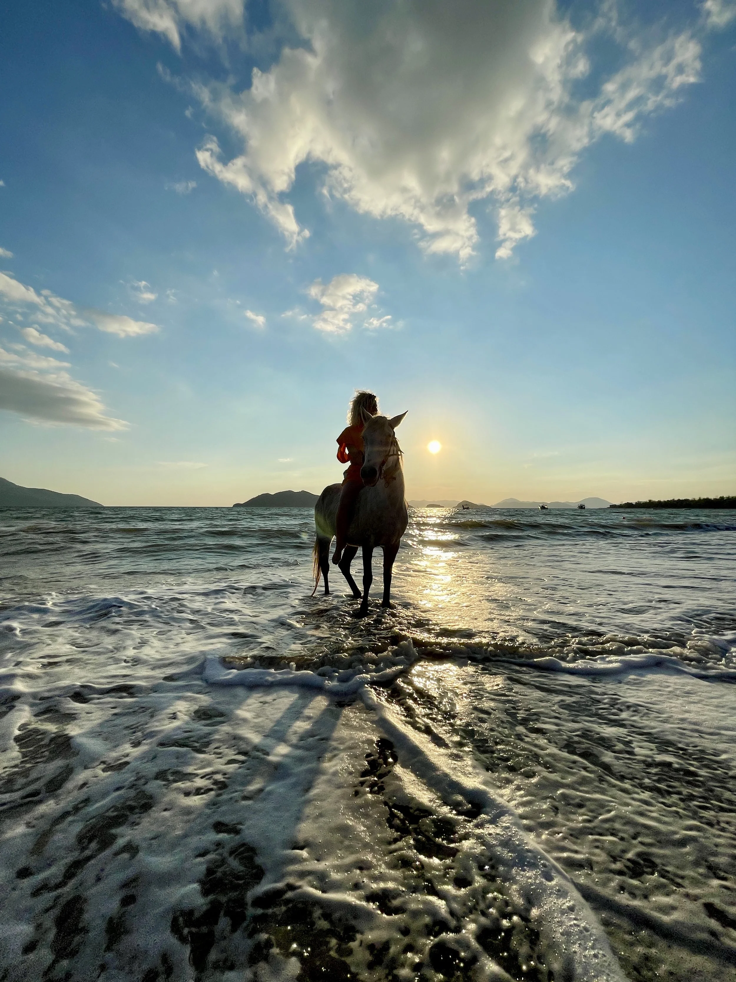 A person riding a horse in the ocean at sunset with a cloudy sky and mountains in the background.