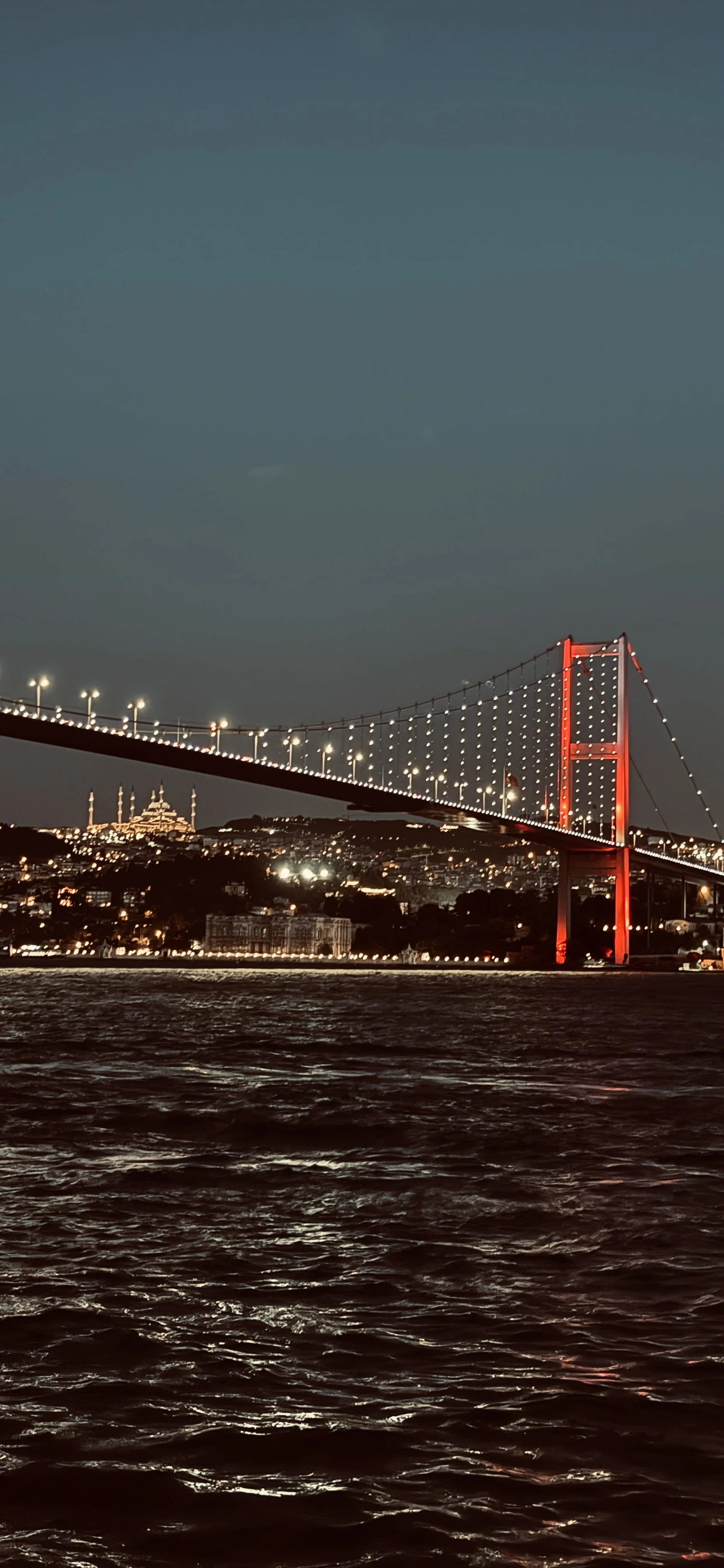 Nighttime view of a illuminated suspension bridge over water, with city lights and a skyline in the background.