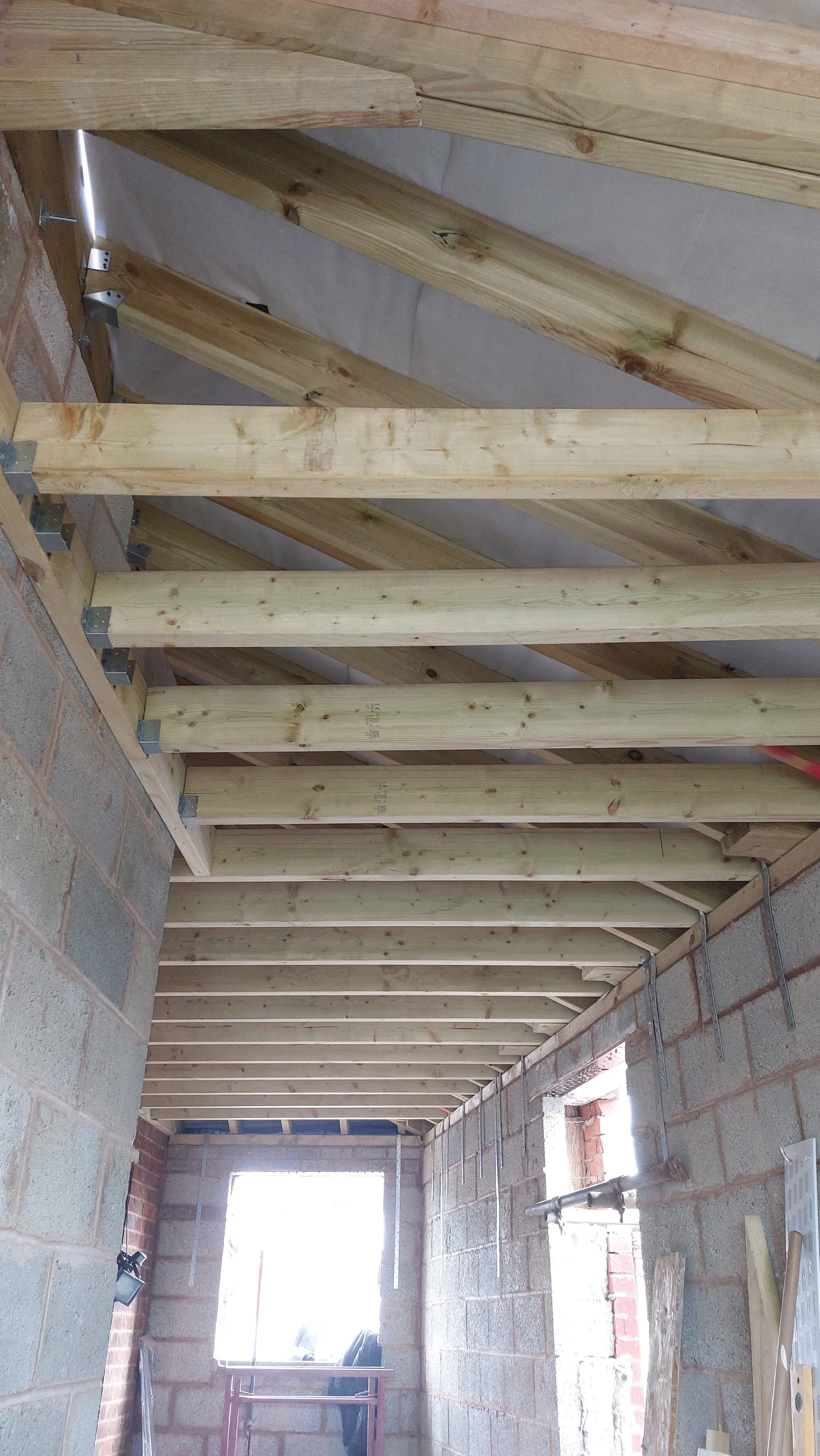 Interior view of a building under construction, showing wooden ceiling beams and joists, with brick and concrete walls, and a window at the end of the space.