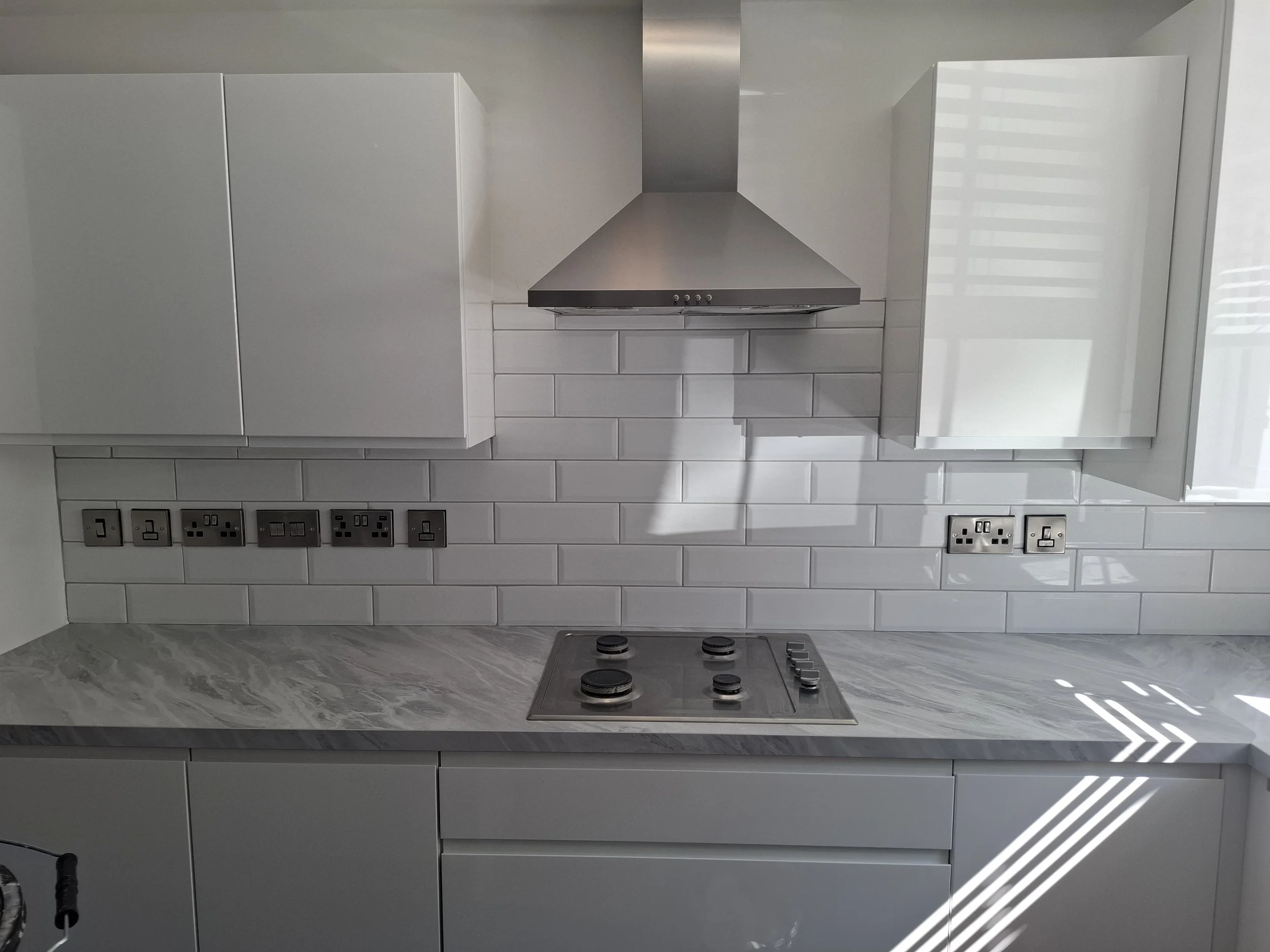 Modern white kitchen with wall-mounted cabinets, gray marble countertop, and a stainless steel stove top with four burners, stainless steel range hood, and white brick-style backsplash. Multiple electrical outlets are visible along the backsplash.