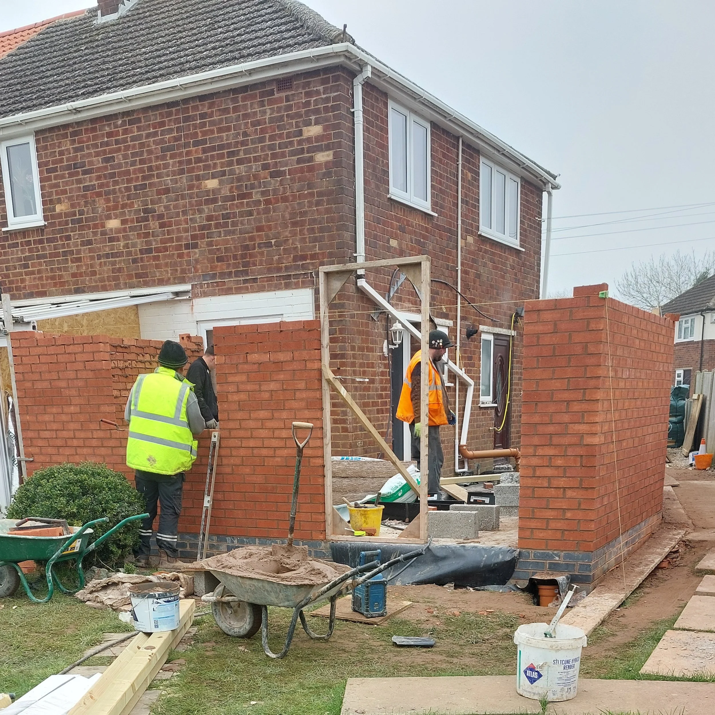 Construction workers building a brick wall on a residential property, with tools and construction materials around.