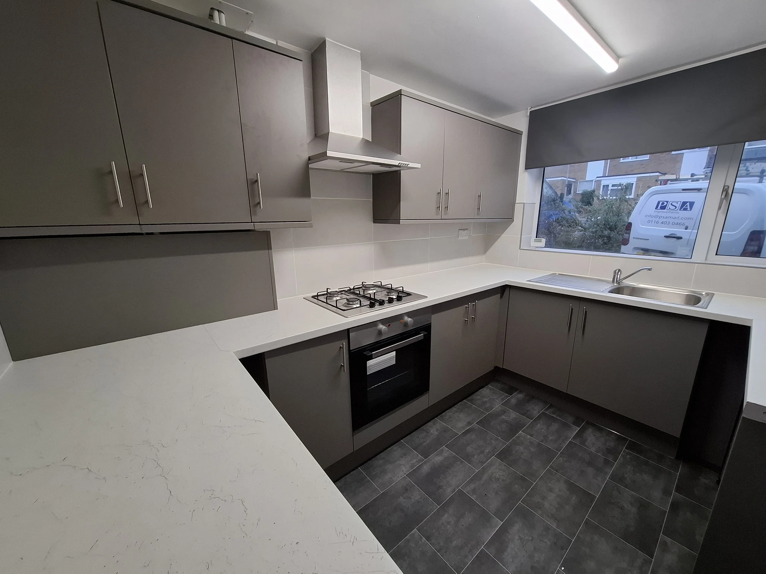 Modern kitchen with gray cabinets, white countertops, a built-in oven, a gas stove, a range hood, a sink near a large window, and gray tiled flooring.