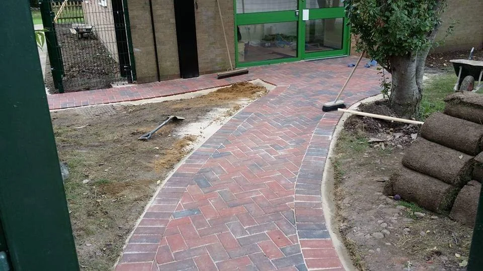 A newly paved brick pathway with curved edges in a backyard, bordered by grass, with a tree on the right. There are gardening tools, including a rake and a shovel, left on the path and nearby. In the background, there is a brick building with a green
