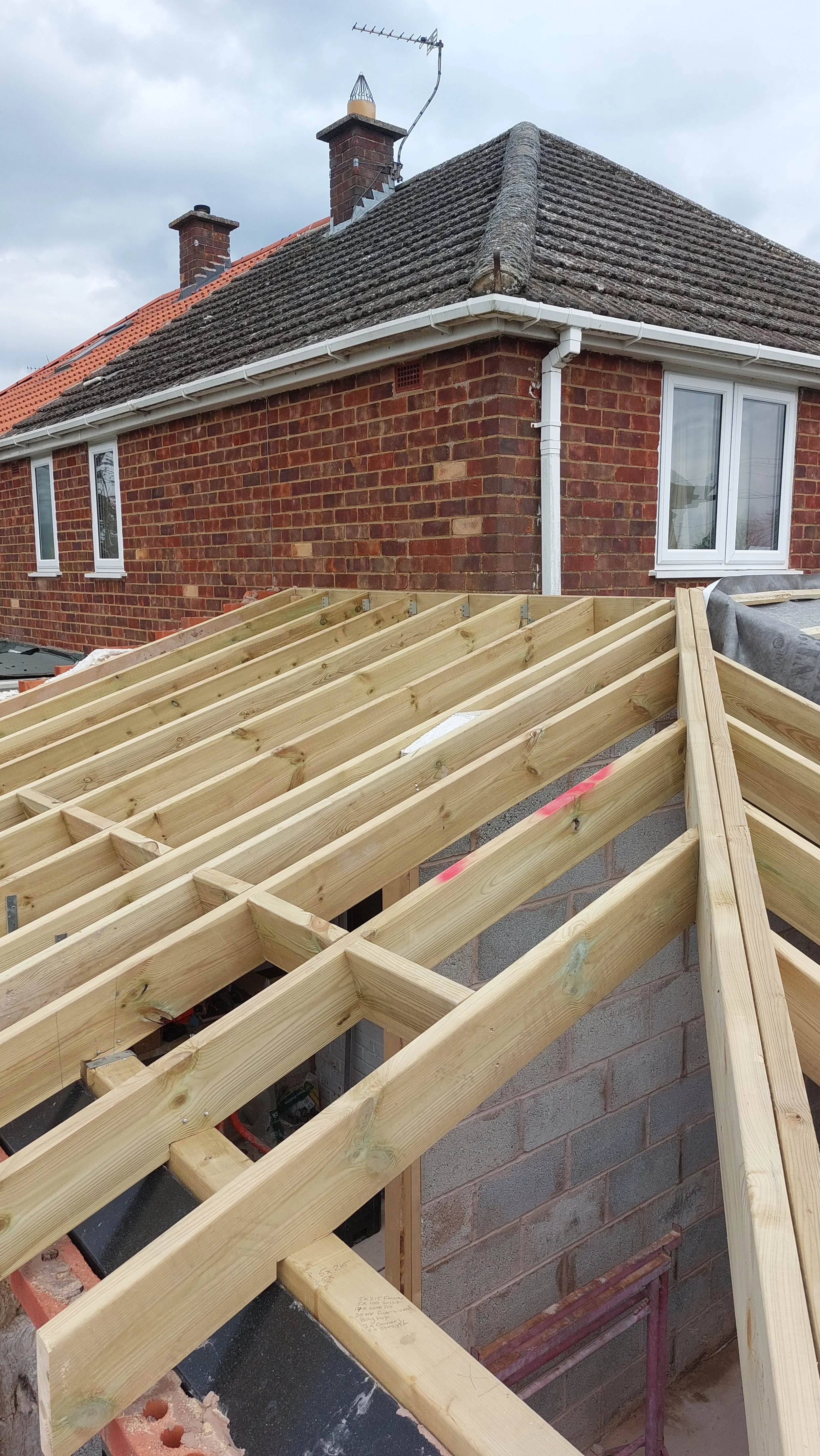 Wooden roof framing construction on a house under construction, with brick wall and upper house with chimney and roofing tiles visible.