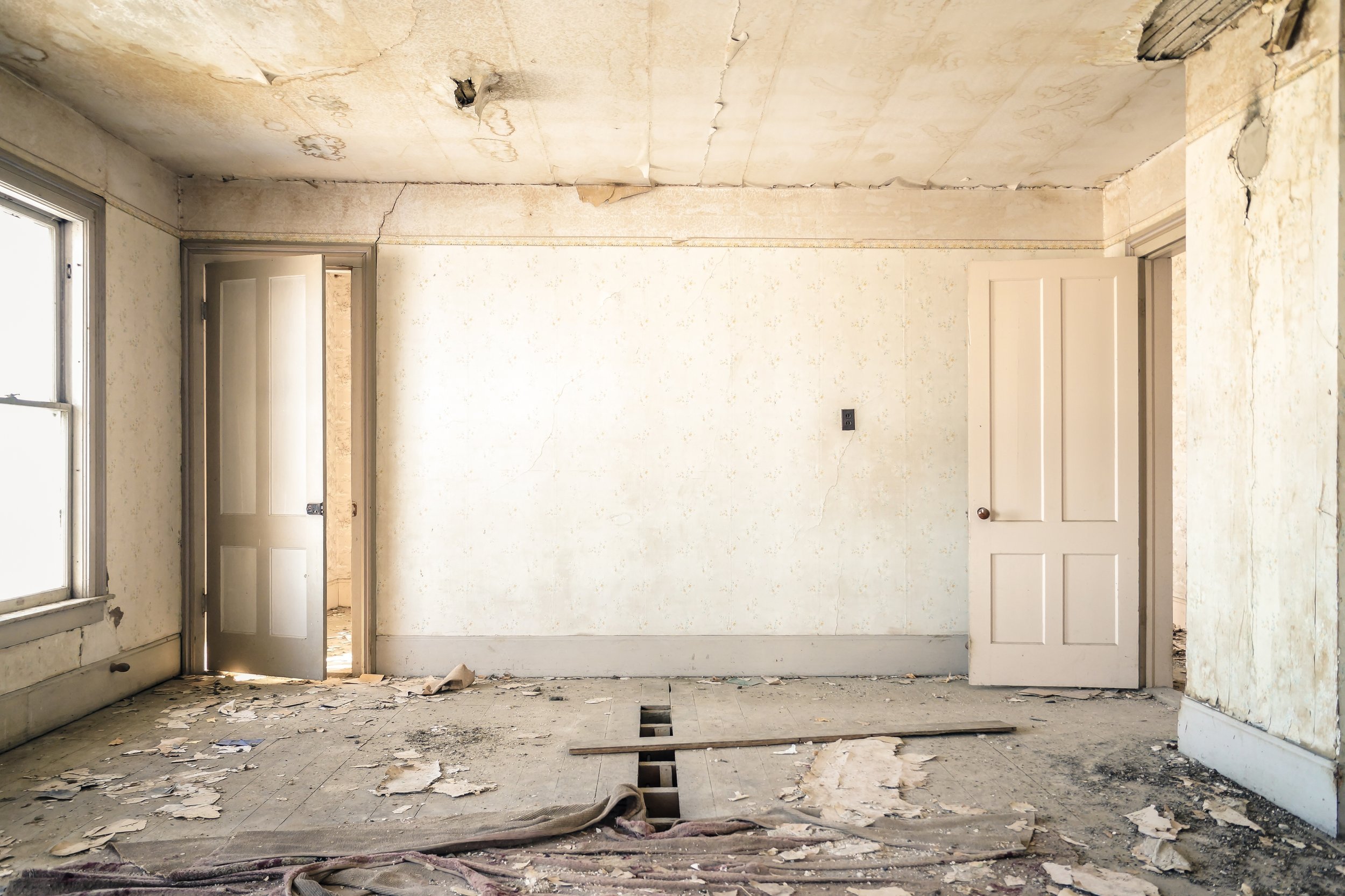 A dilapidated room with peeling wallpaper, a damaged ceiling with a hole, and a partially removed floor revealing underneath construction. There are two doors and a window, all showing signs of neglect.