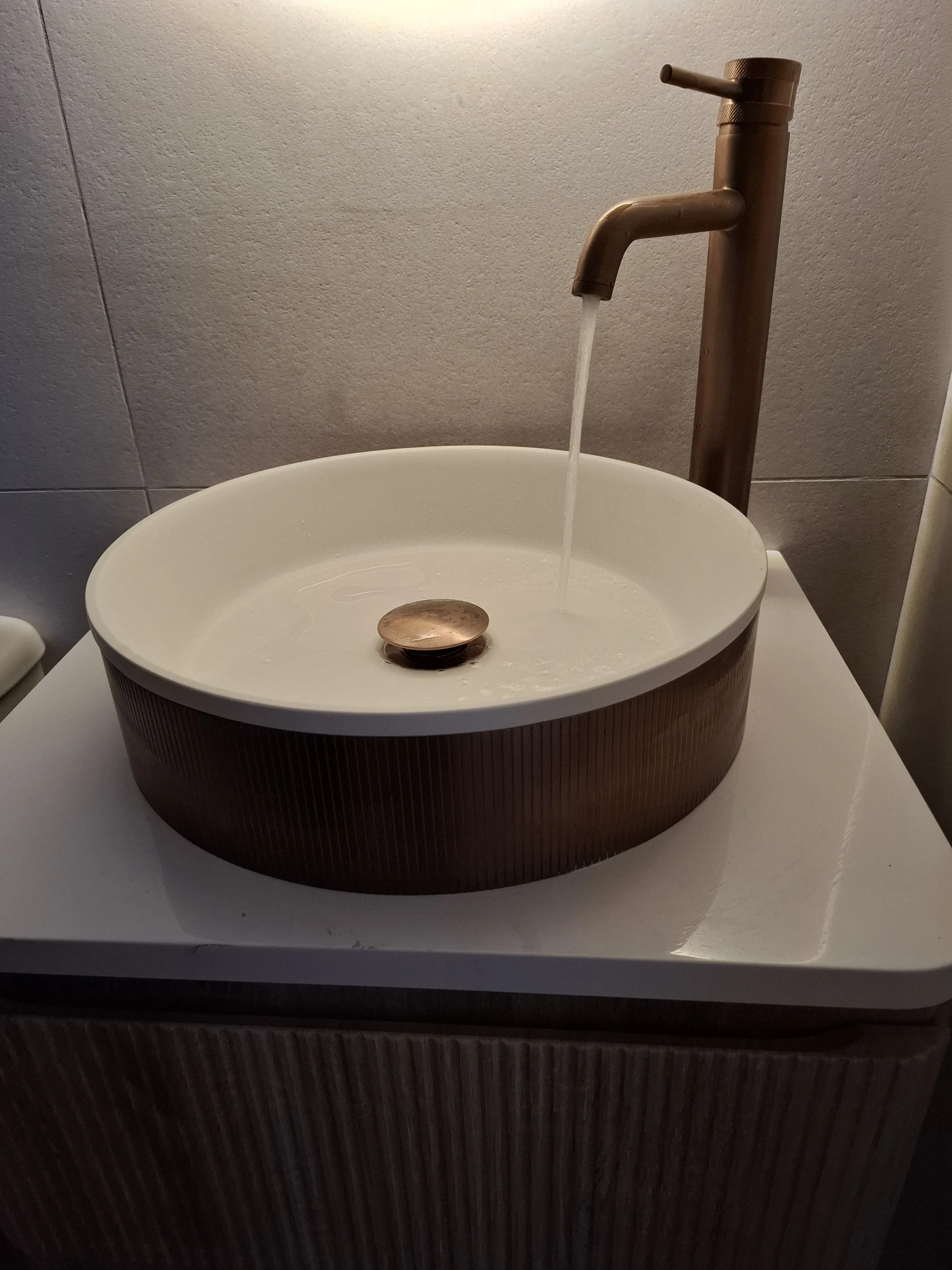 A sink with a bronze faucet and a copper circular drain cover, positioned on a white countertop against a tiled wall.