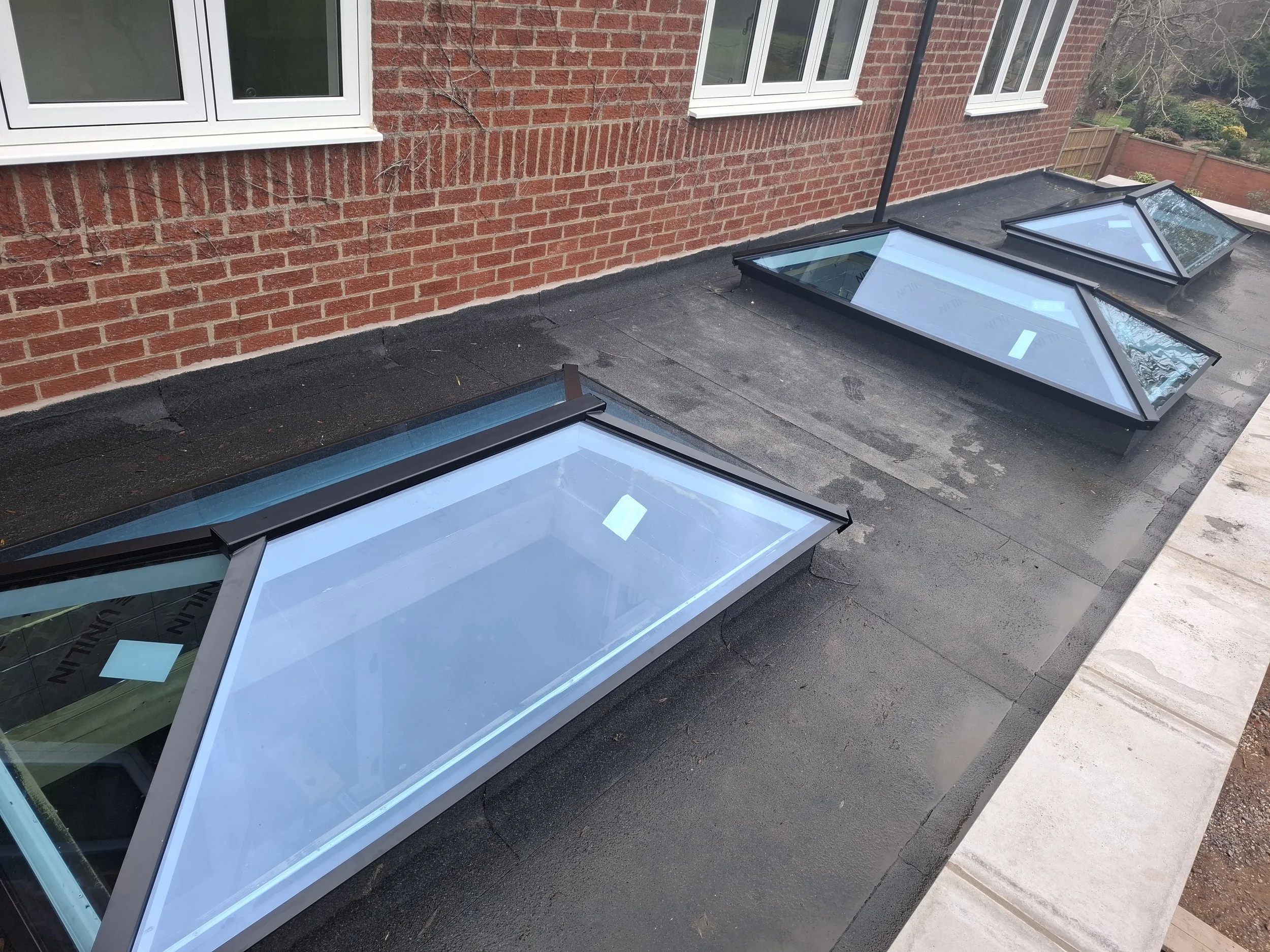 Three large triangular skylights installed on a flat rooftop with a red brick building wall and white-framed windows in the background.