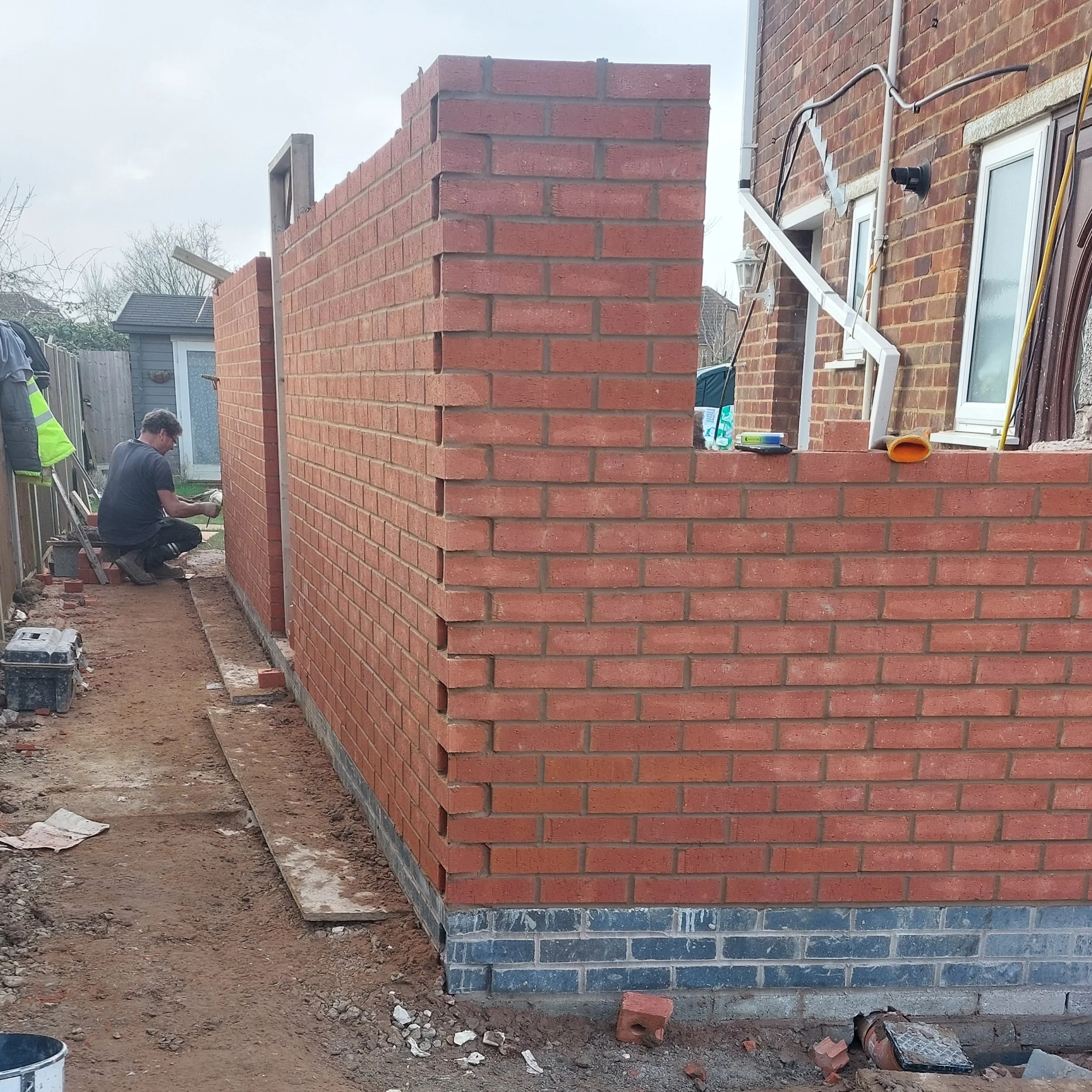 A construction worker laying bricks to build an exterior wall of a building, with tools and supplies nearby.