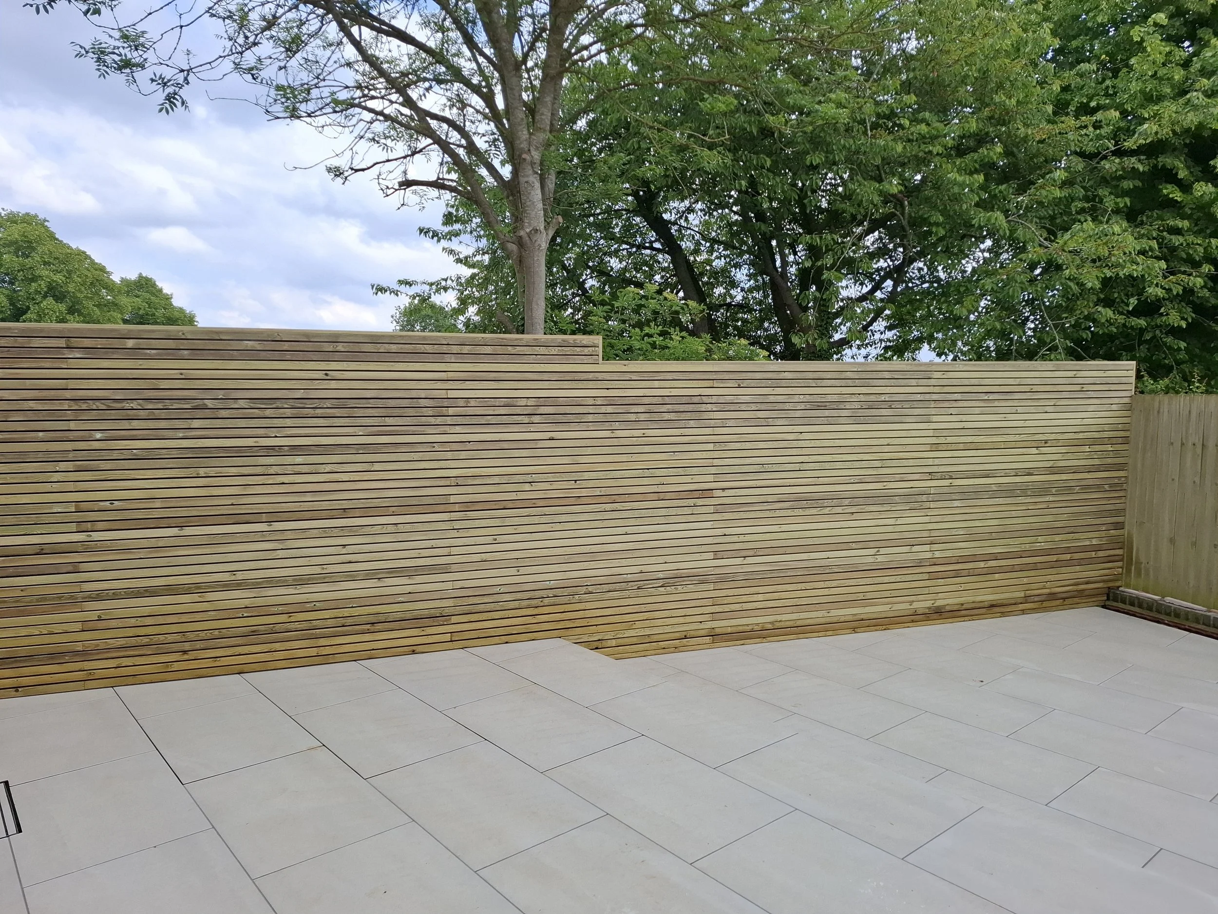 A backyard patio with large beige tiles, surrounded by a wooden slat fence and green trees in the background under a partly cloudy sky.