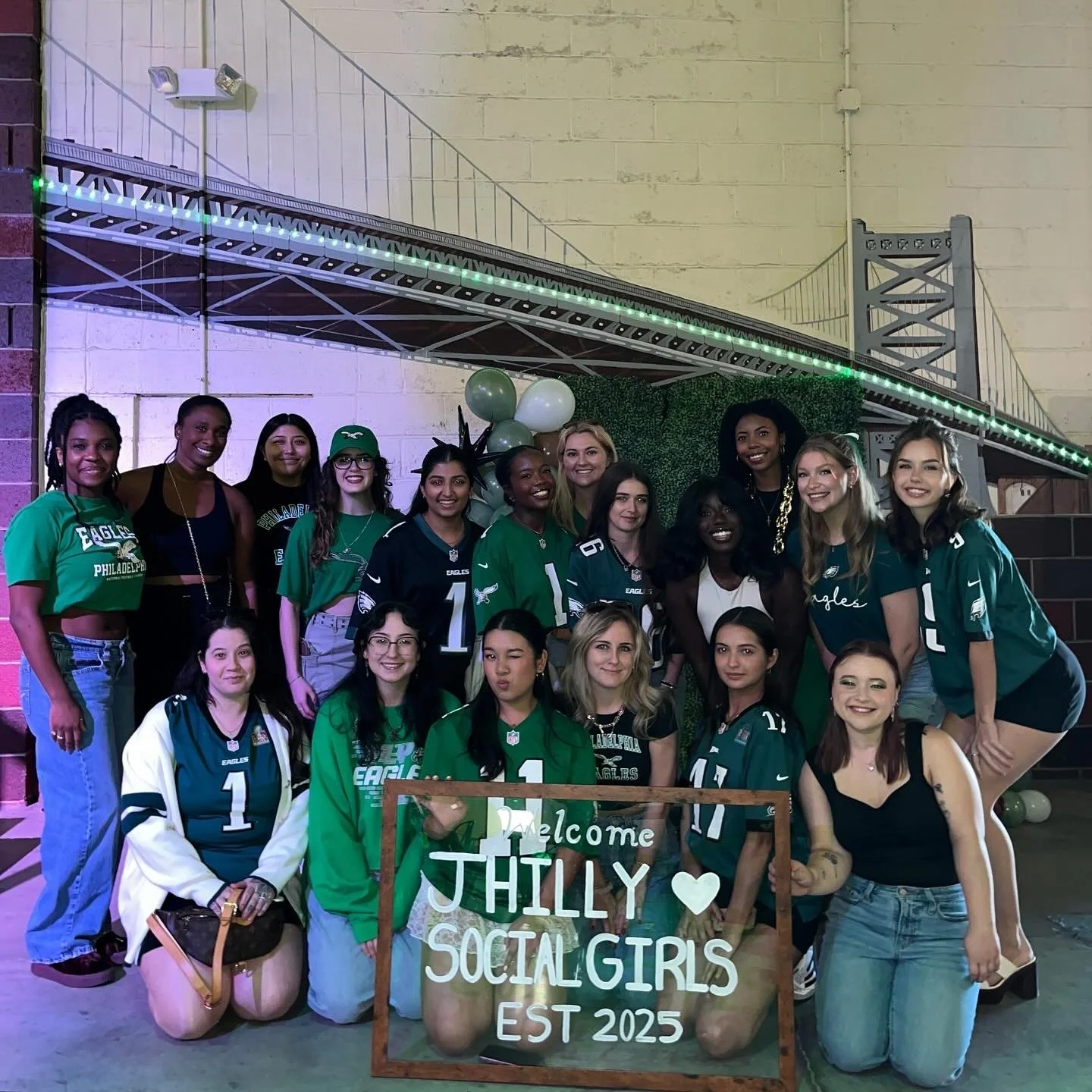 Group of young women celebrating at a social gathering, some wearing Philadelphia Eagles jerseys, holding a wooden frame with a sign that reads 'Welcome Thilly Social Girls Est 2025', in front of a decorative backdrop of balloons and greenery, with a bridge mural on the wall behind.