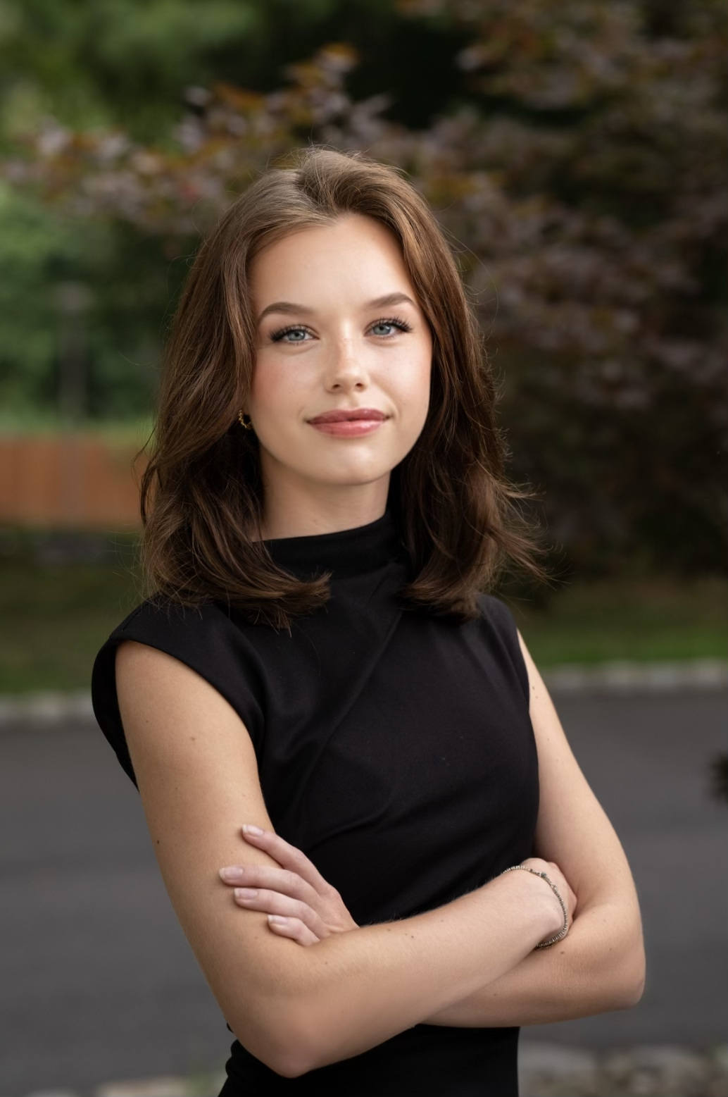 A young woman with shoulder-length brown hair, wearing a black sleeveless top, standing outdoors with crossed arms, smiling softly at the camera, with trees and greenery in the blurred background.