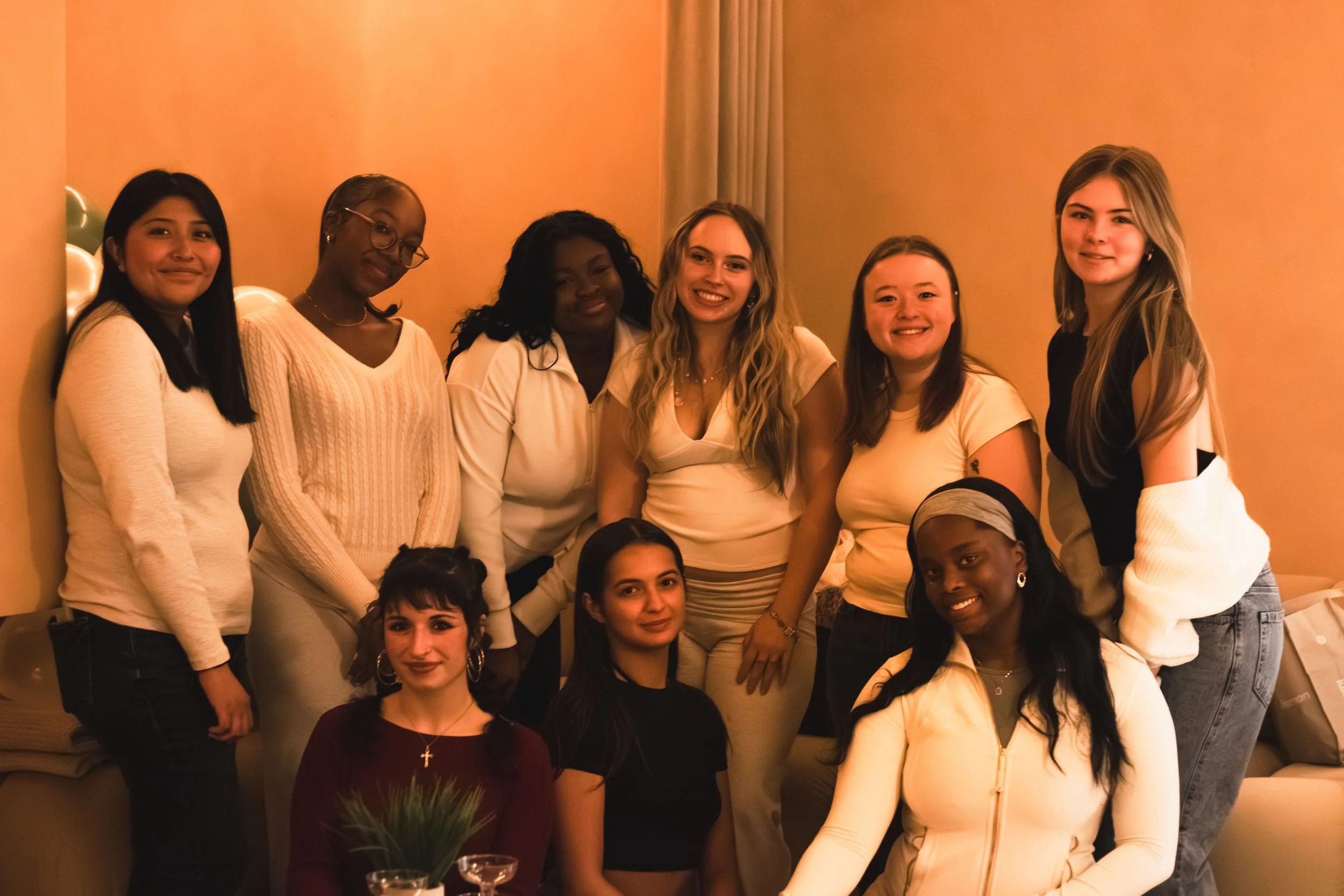 Group of nine women gathered indoors, posing for a photo, smiling, with warm lighting and balloons in background.
