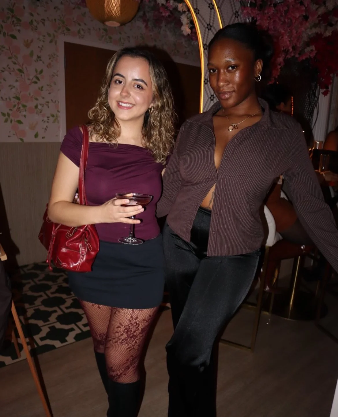 Two women posing together at a social gathering; one holding a drink in a glass.