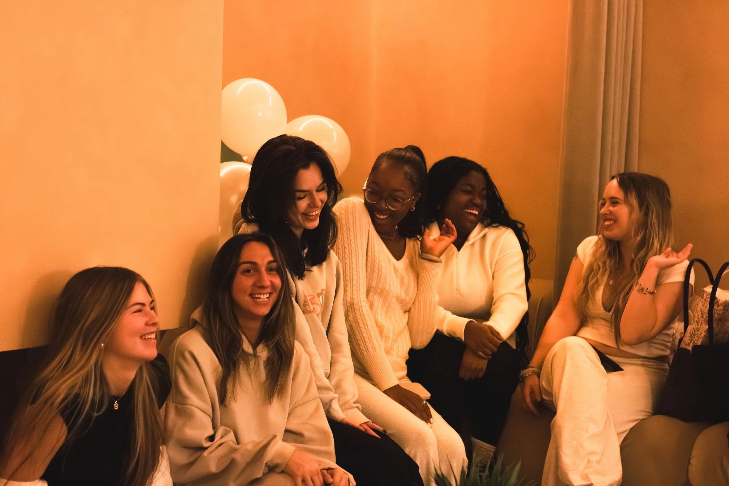 Group of six women sitting on a couch and laughing together at a birthday celebration with balloons in the background.