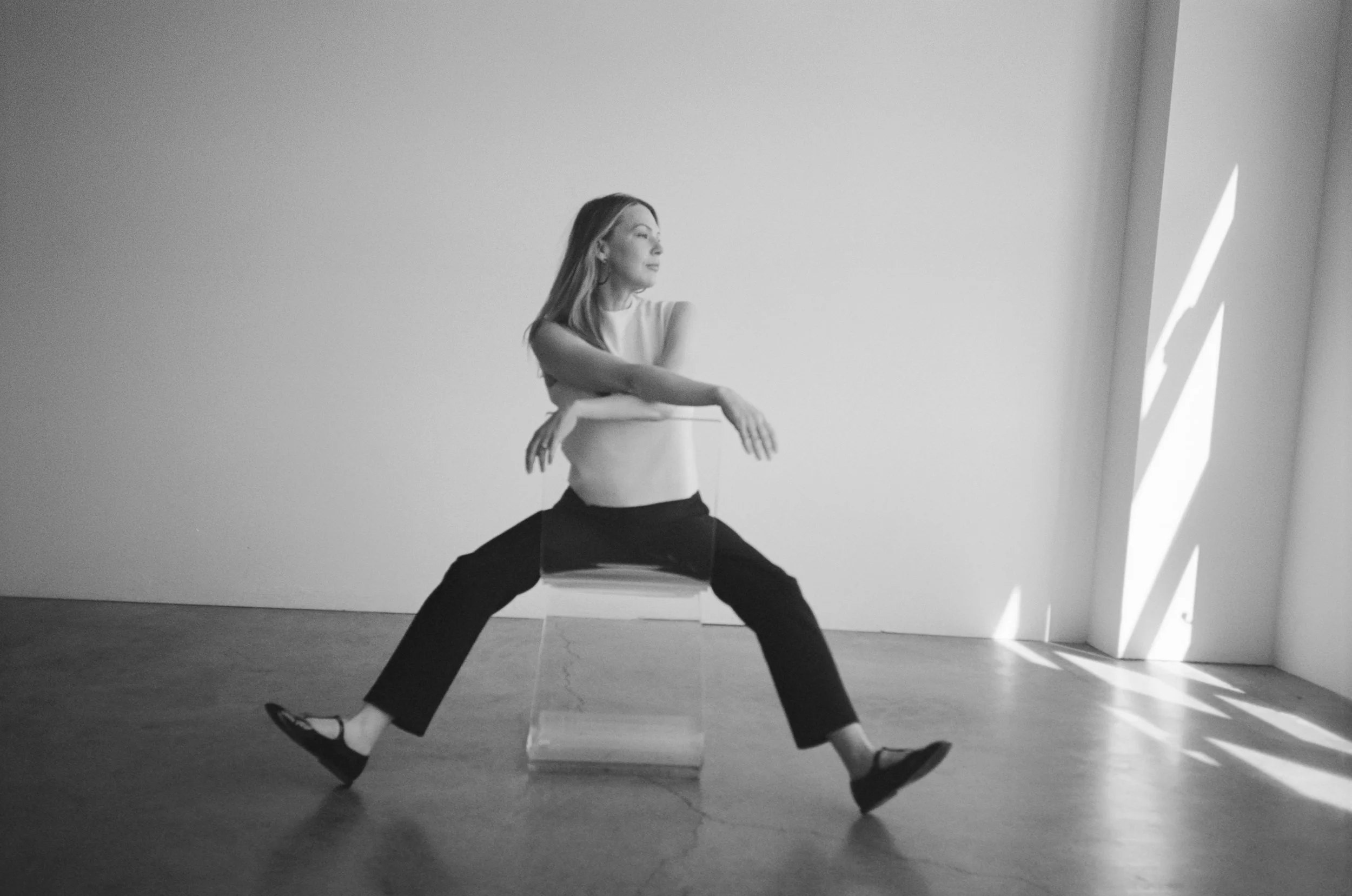 A woman in a white top and black pants demonstrating a dance pose in a minimalist room with light coming through vertical slats.