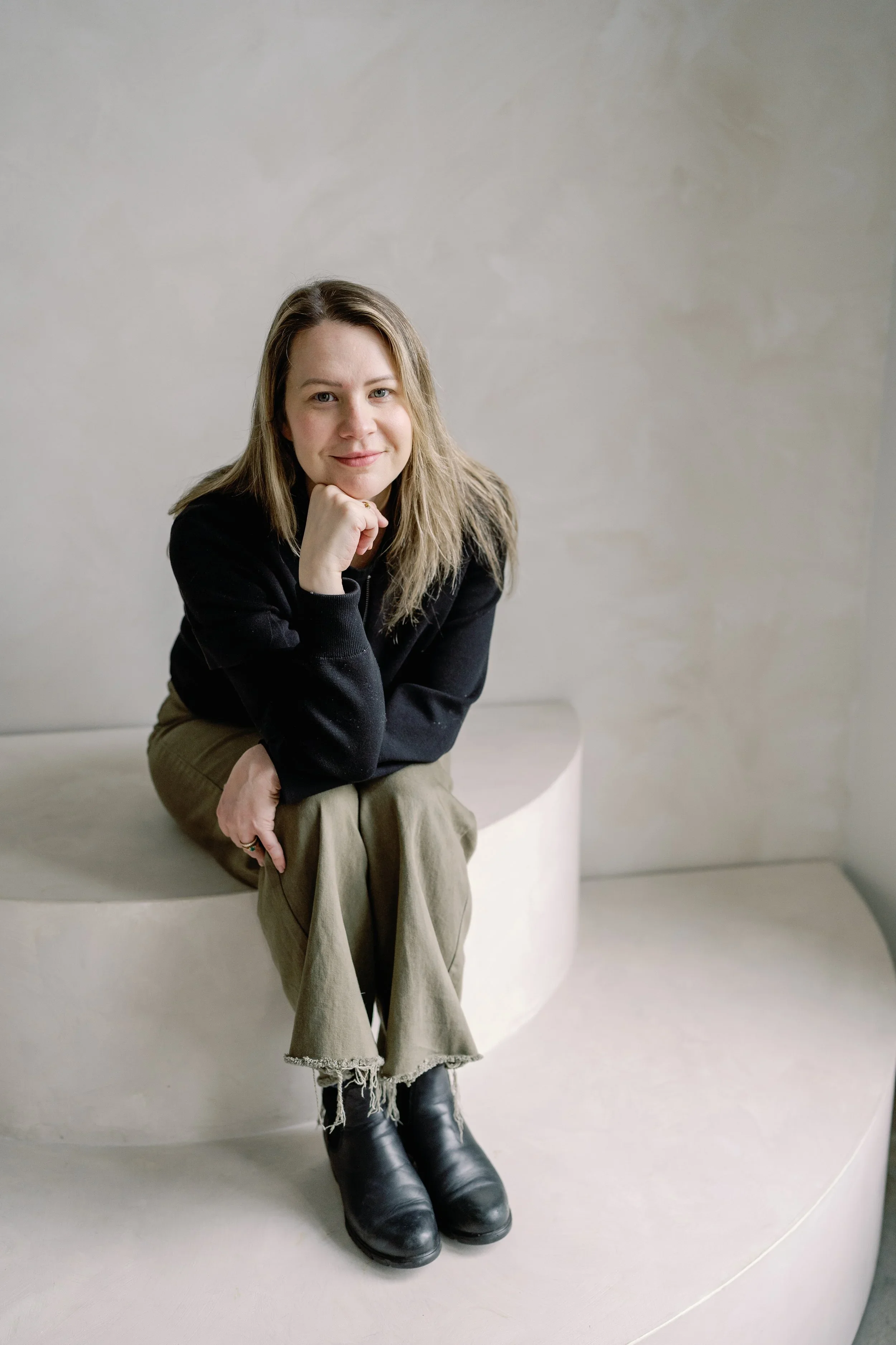 A woman with blonde hair, wearing a black top, tan pants with frayed hems, and black boots, sitting on a curved bench or surface against a plain, light-colored wall, resting her chin on her hand.