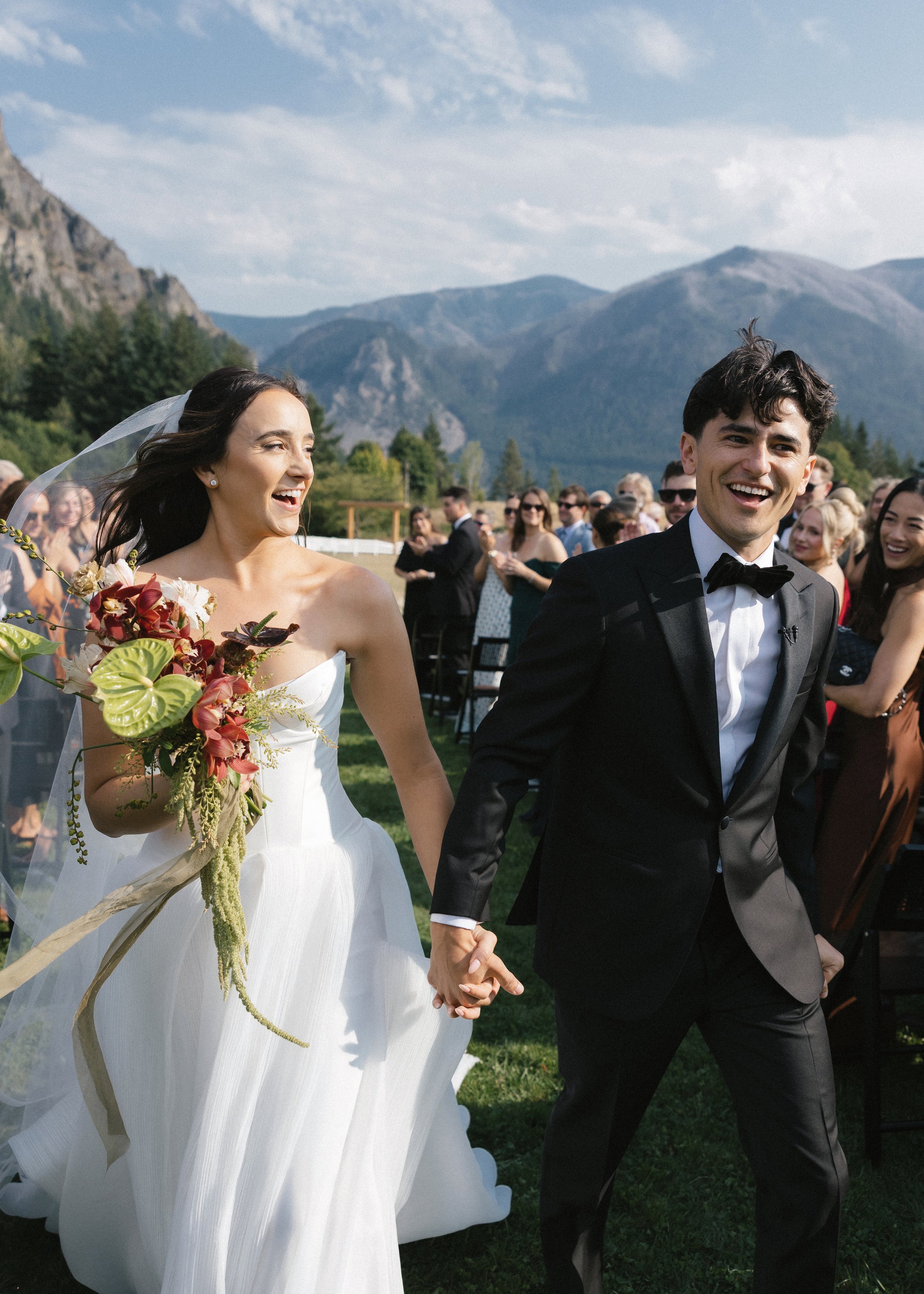 A bride and groom holding hands at an outdoor wedding ceremony, with guests in the background and mountains in the distance under a partly cloudy sky.