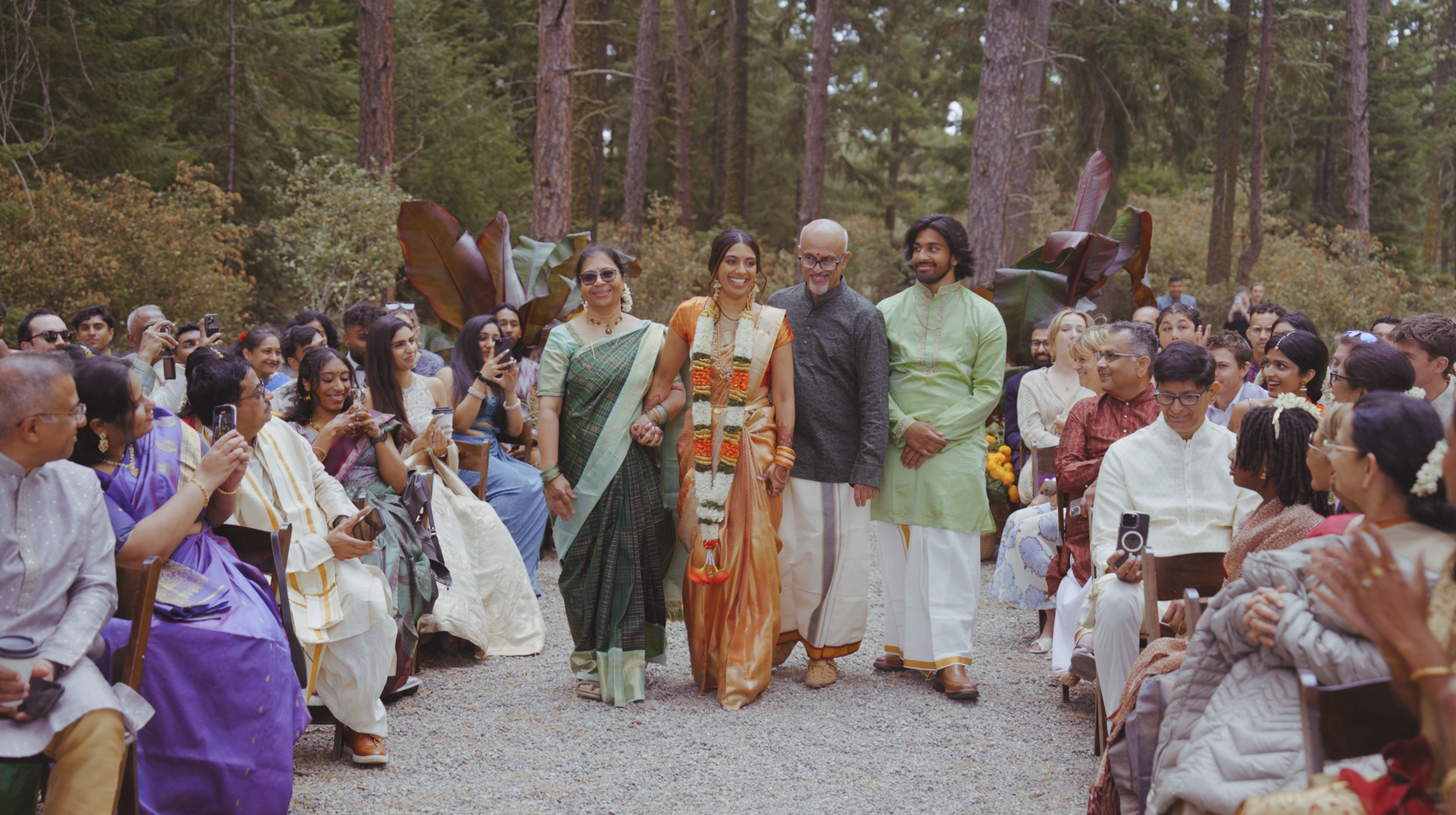 Group of people gathered outdoors in a wooded area for a cultural celebration; some are taking photos, and a couple of individuals are walking down the central aisle, including a woman in an orange sari and a man wearing a garland from their necks.