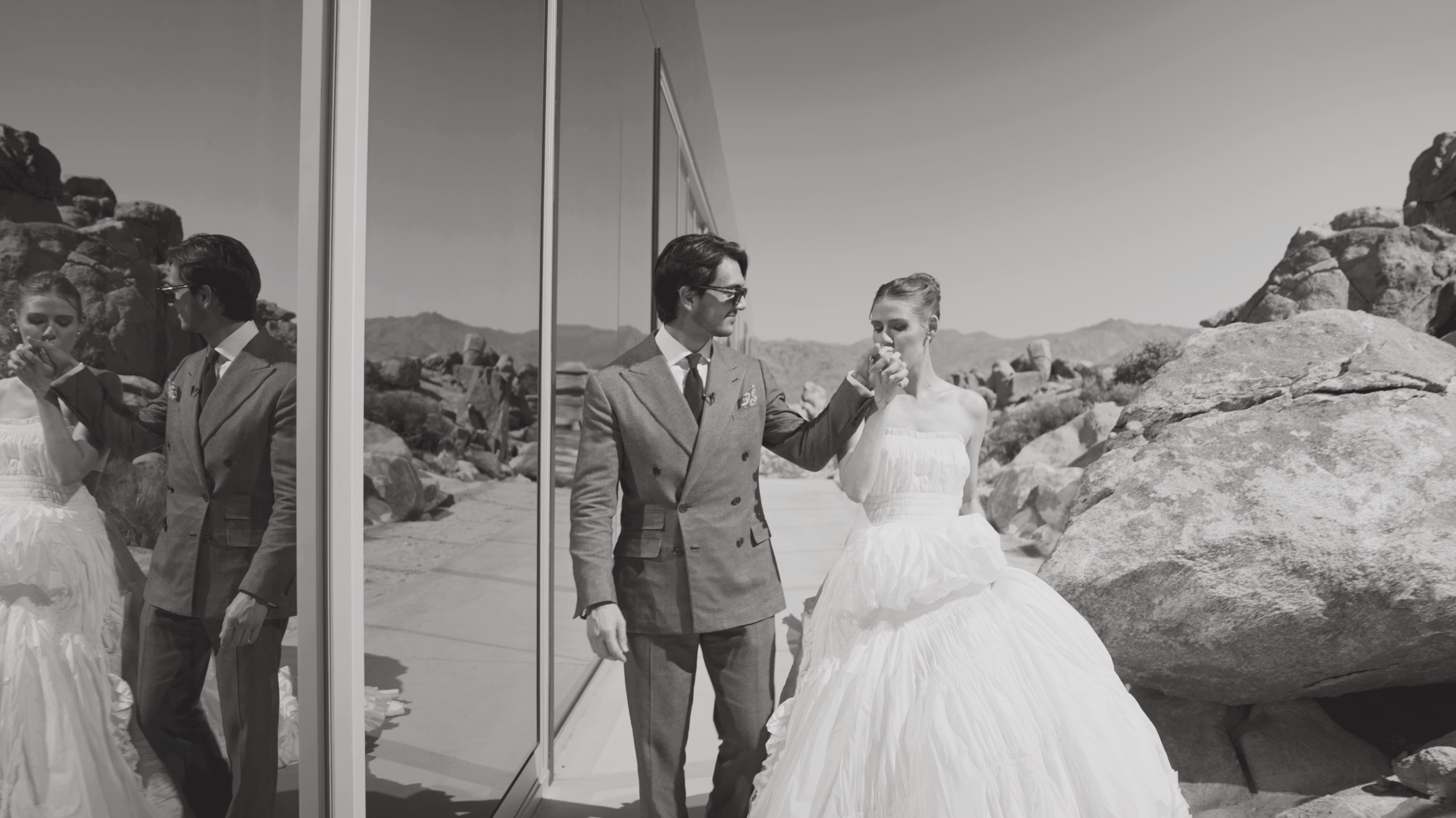 A man in a suit and glasses gently touches the hand of a woman in a strapless, ruffled white dress, with rocky desert landscape and mountains in the background.