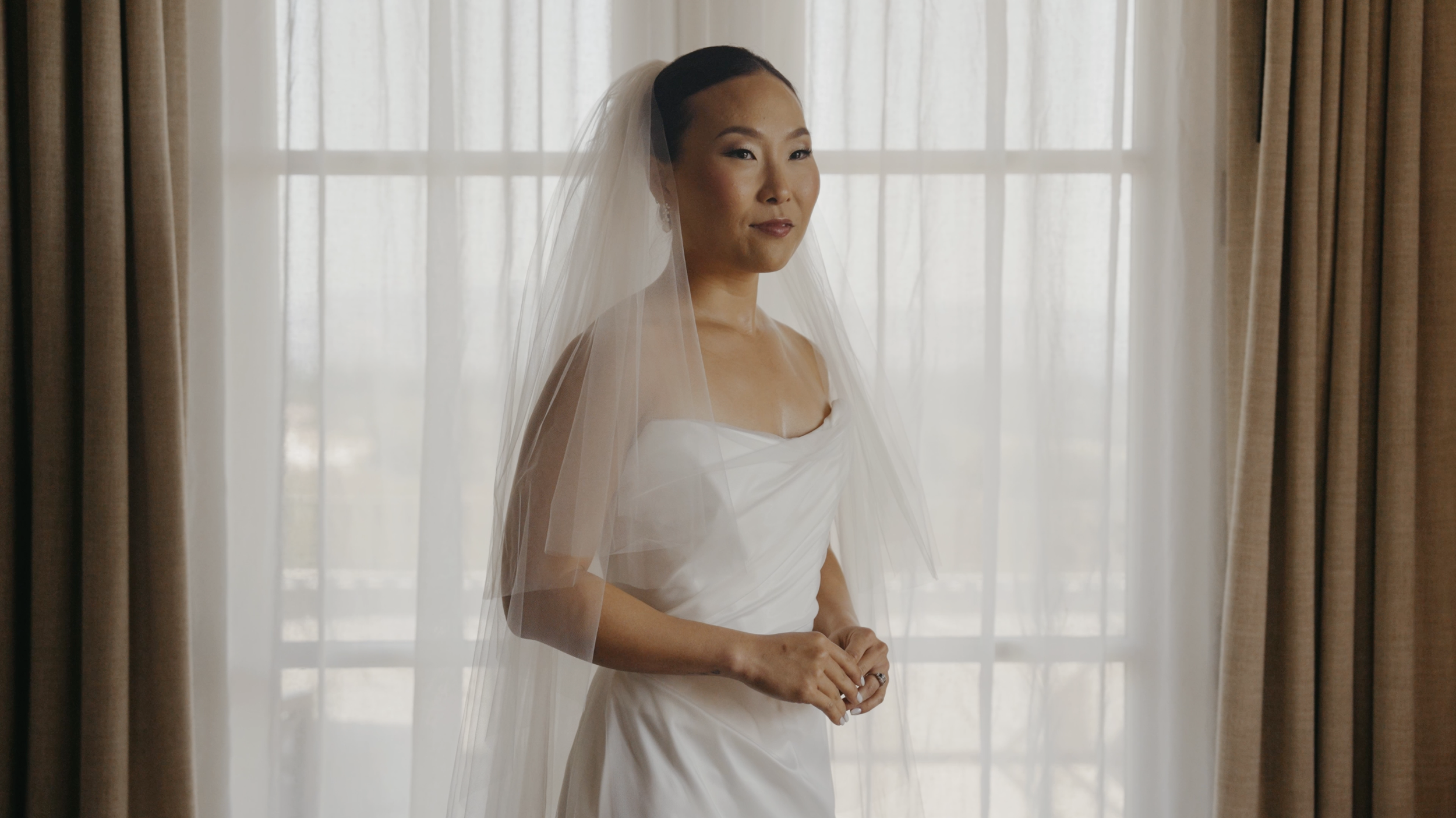 A bride in a white dress with a veil standing in front of a window with sheer curtains.