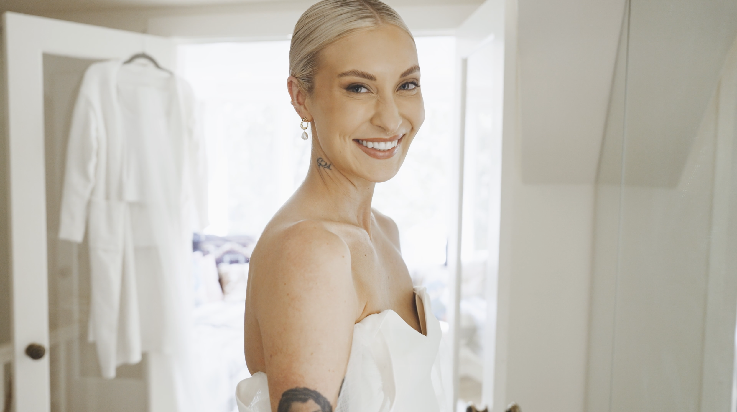 A smiling woman with blonde hair styled slicked back, showing her right shoulder and a tattoo on her neck, in a room with white walls and a white dress hanging in the background.