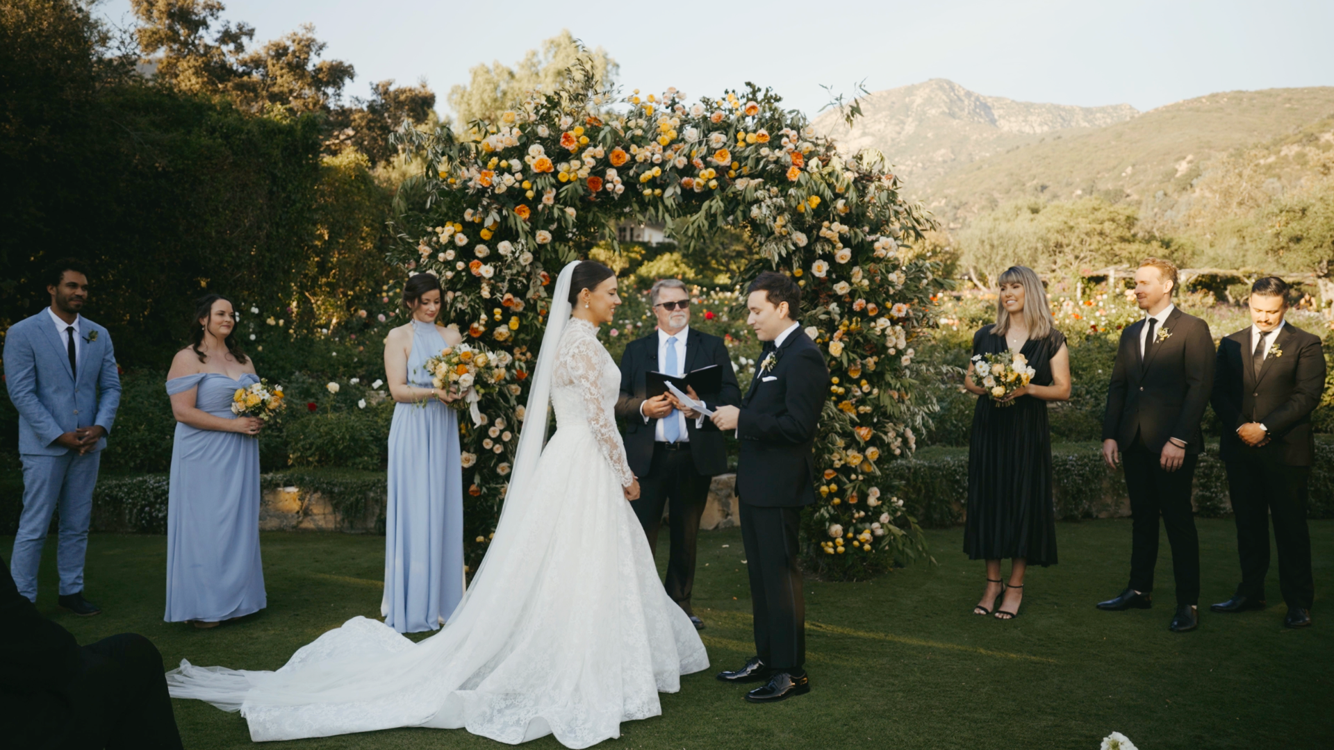 A wedding ceremony taking place outdoors with a bride and groom standing under a floral arch surrounded by bridesmaids and groomsmen.