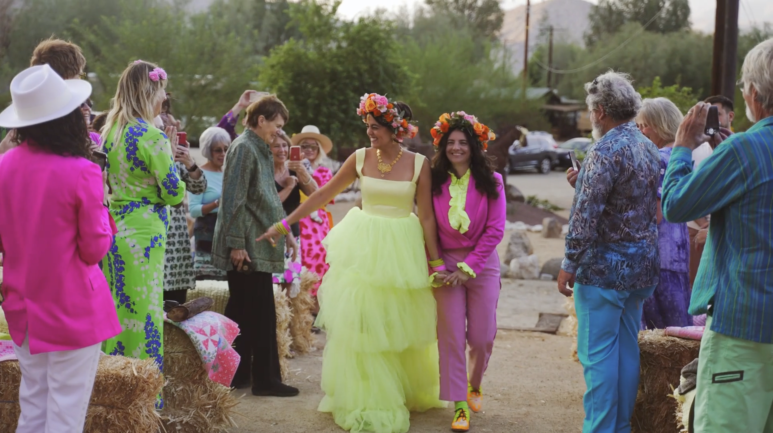 People gathering outdoors in colorful outfits, with two women wearing flower crowns and bright dresses smiling at each other, surrounded by onlookers taking photos.