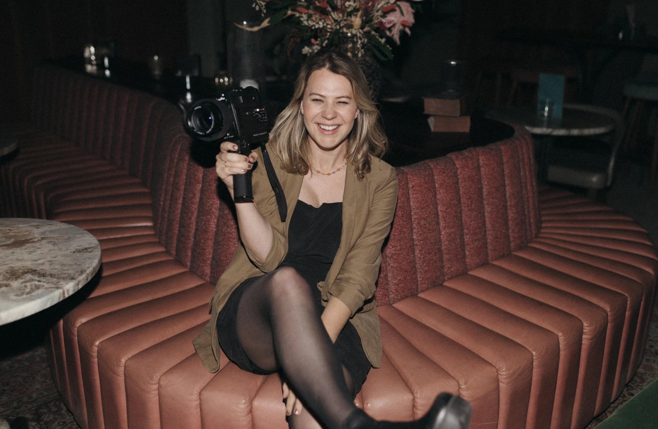 A woman with blonde hair, smiling and squinting, sitting on a pinkish-red booth in a dimly lit venue, holding a camera in her right hand. She is wearing a black dress with sheer black tights and an olive green jacket. A floral arrangement is visible in the background.