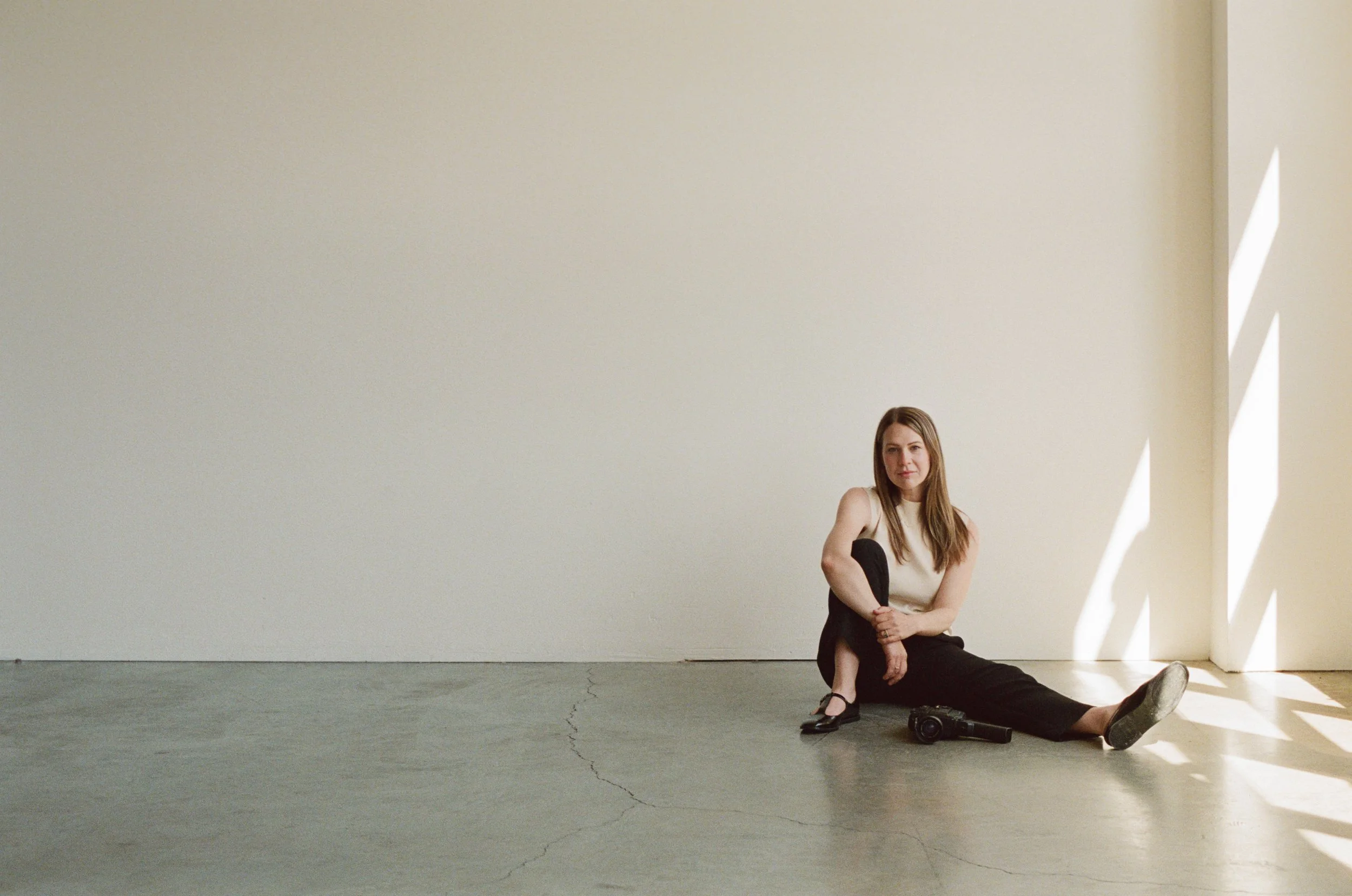 Woman with brown hair sitting on the floor, leaning against a white wall with sunlight streaming through window blinds, holding a camera, wearing black pants, a sleeveless top, and heels.