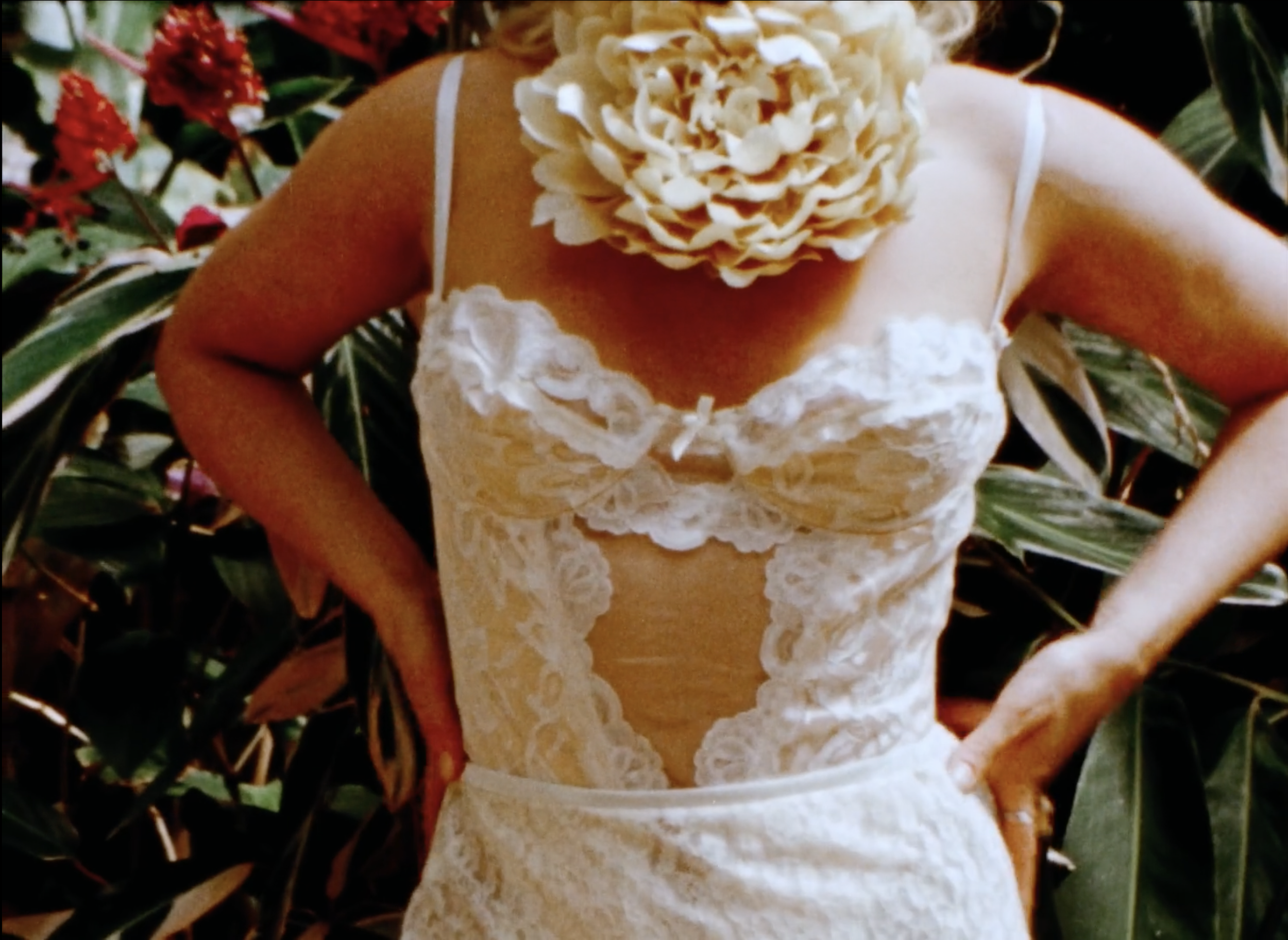 Close-up of a person wearing a white lace dress with floral decorations, standing outdoors among green foliage and red flowers.