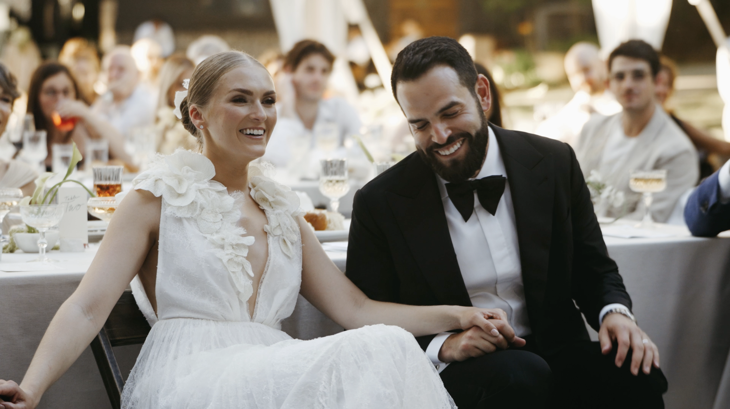 A bride and groom sitting together at their wedding reception, smiling and holding hands, surrounded by guests at decorated tables outdoors.