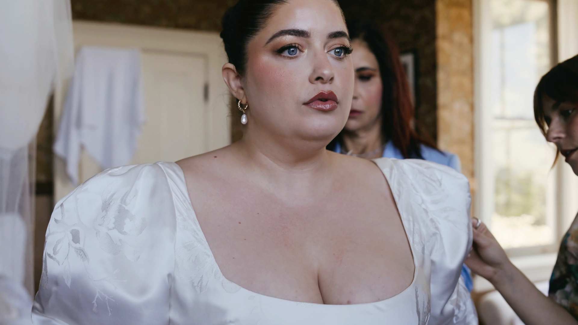 A woman with blue eyes and dark hair in an updo, wearing pearl earrings and a white satin dress with puffed sleeves, looking serious. Two other women are in the background.