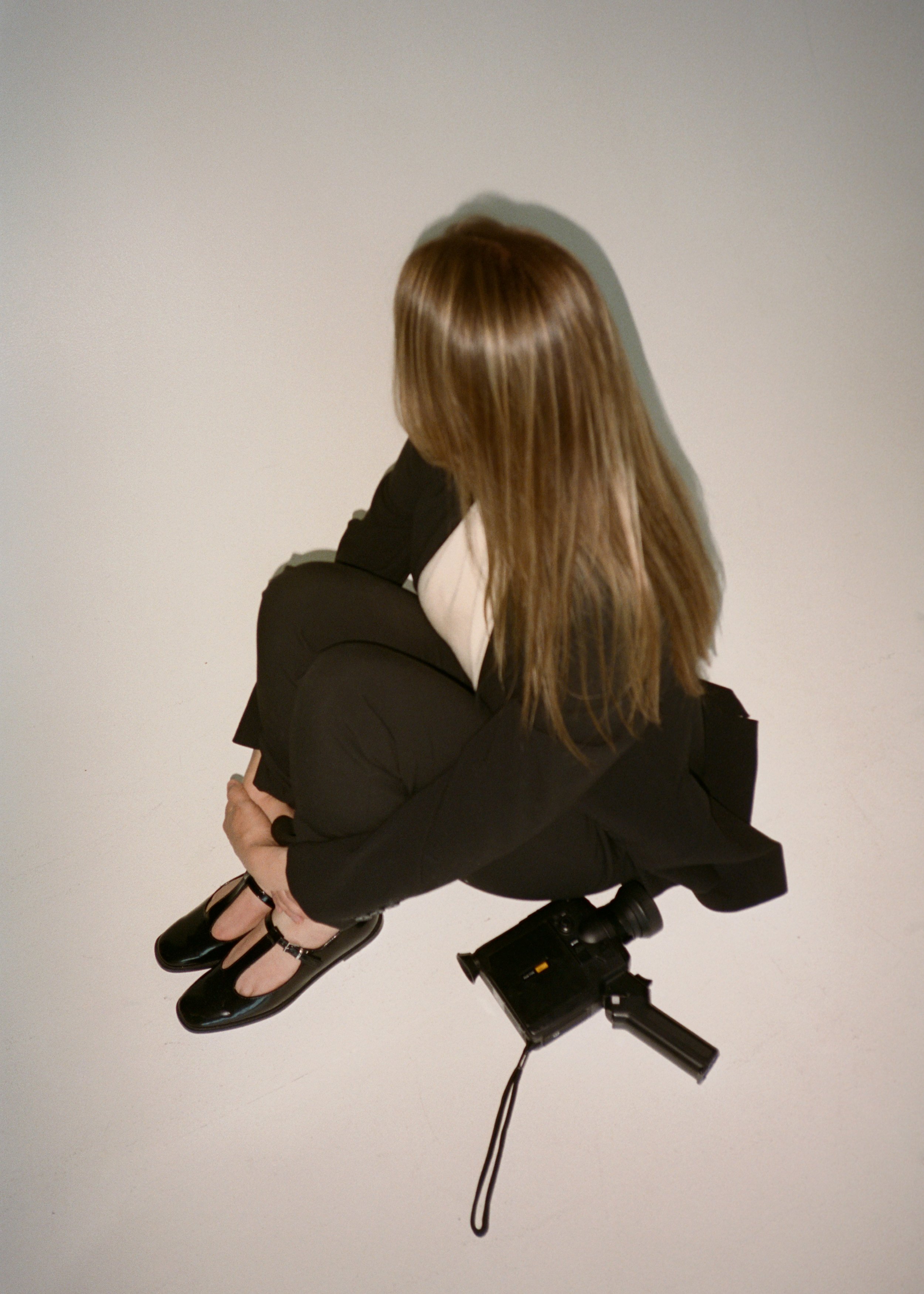 A person with long brown hair, dressed in a black jacket, white shirt, and black shoes, sitting on a stool with a camera mounted on a tripod in front. The person is facing away from the camera, with arms crossed and head turned slightly to the right.
