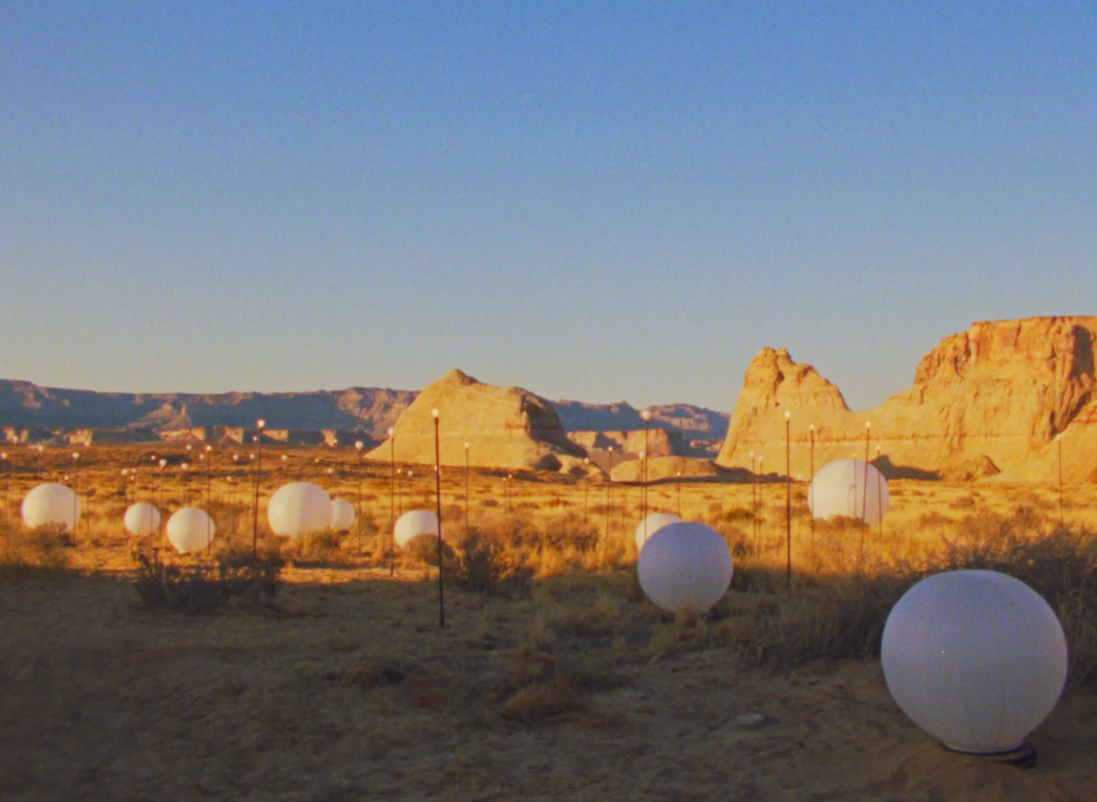 A desert landscape at sunset with large, white spherical objects on the ground and mesas in the background.