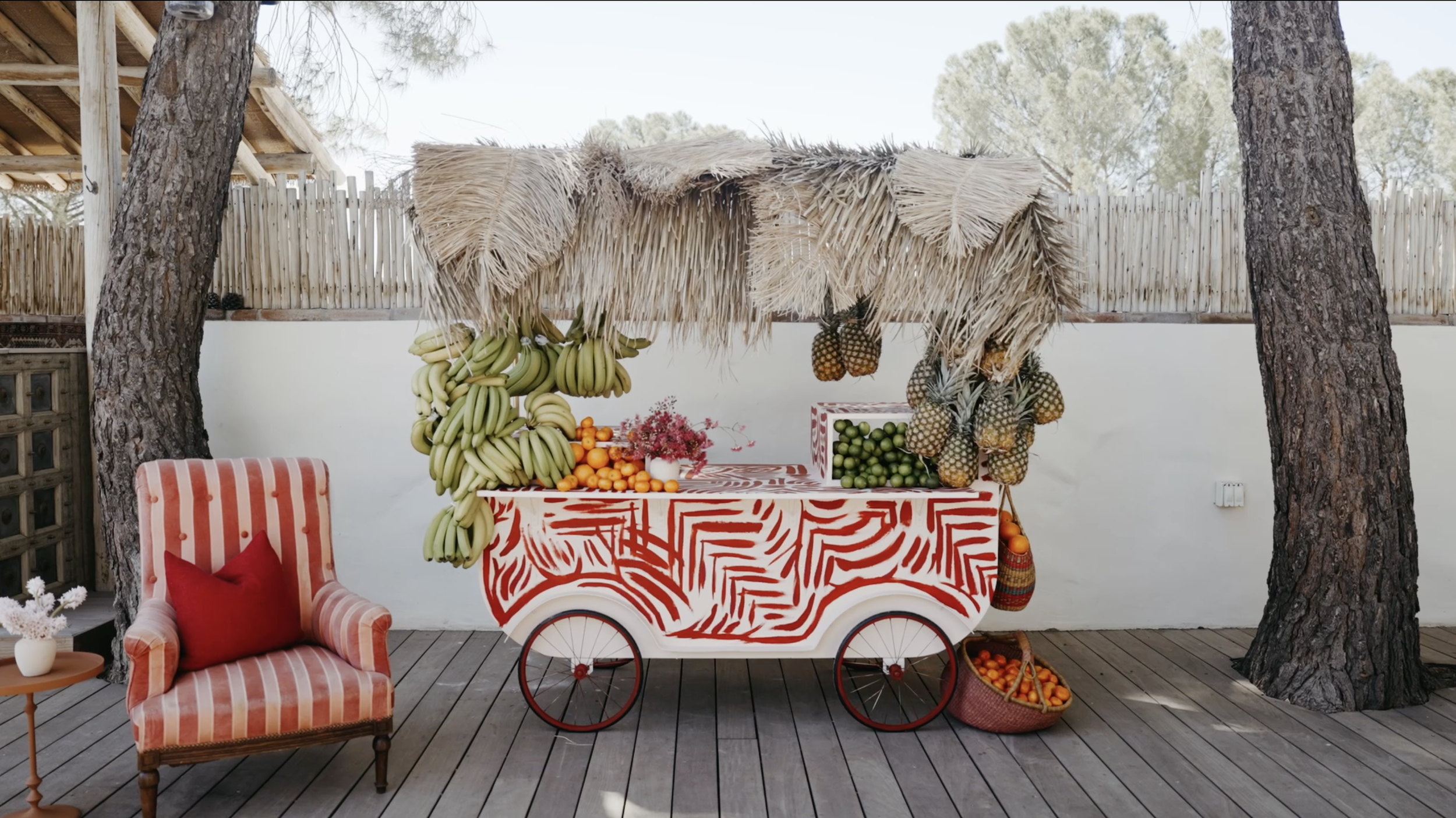 A colorful fruit cart with bananas, oranges, pineapples, and limes, set outdoors among trees, with a red and white patterned table, a striped armchair with a red pillow, and a basket of oranges nearby.