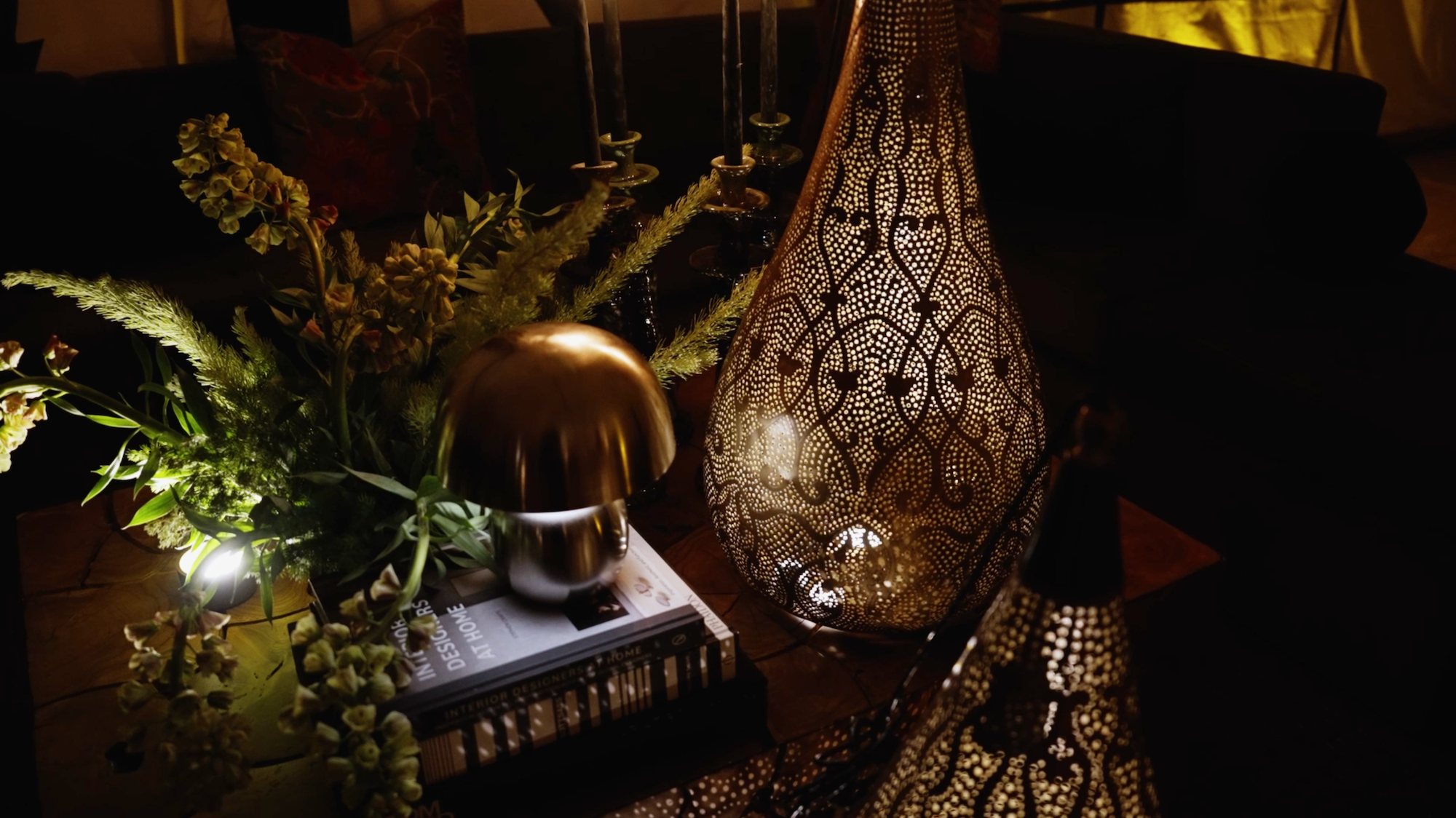 Dimly lit living room or sitting area with a decorative, perforated metal lamp, a flower arrangement, a metallic table lamp, books, and a dark sofa in the background.