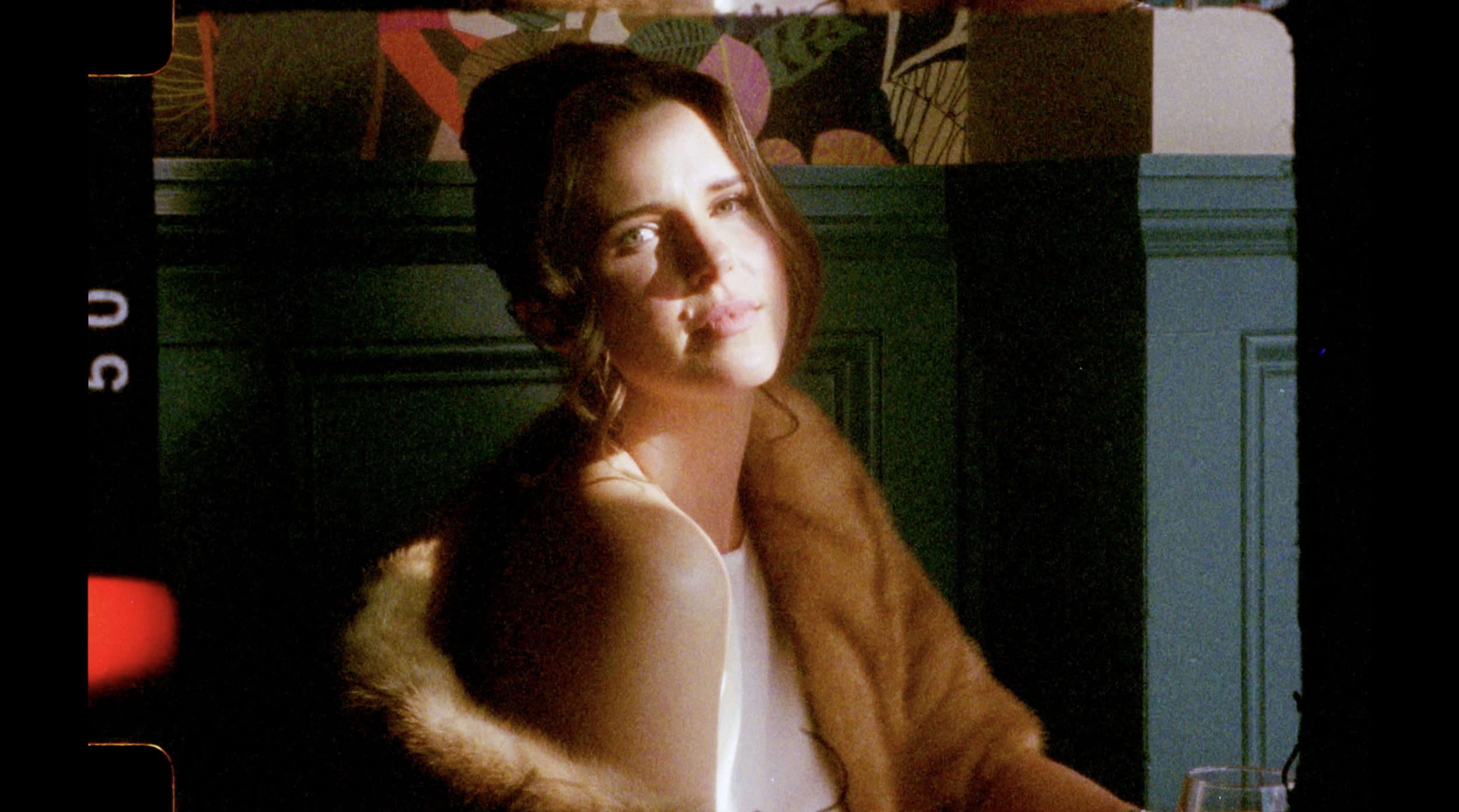 A woman with reddish-brown hair and fair skin sitting on a chair, wearing a white top and a brown fur coat, in a dimly lit room with colorful artwork on the wall behind her.