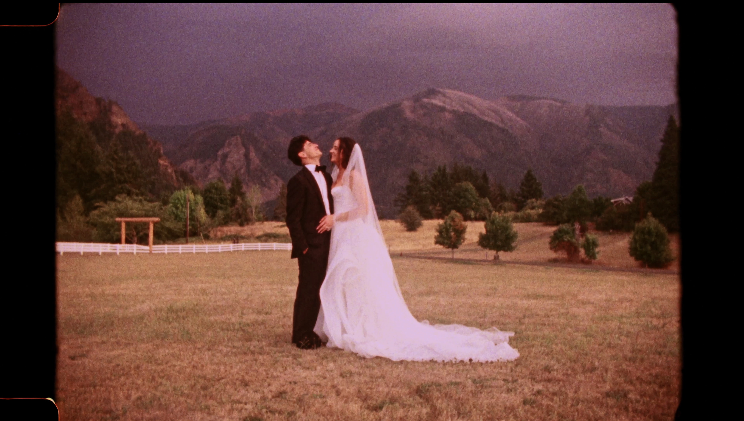 A bride and groom standing outdoors in a field with mountains in the background, celebrating their wedding.