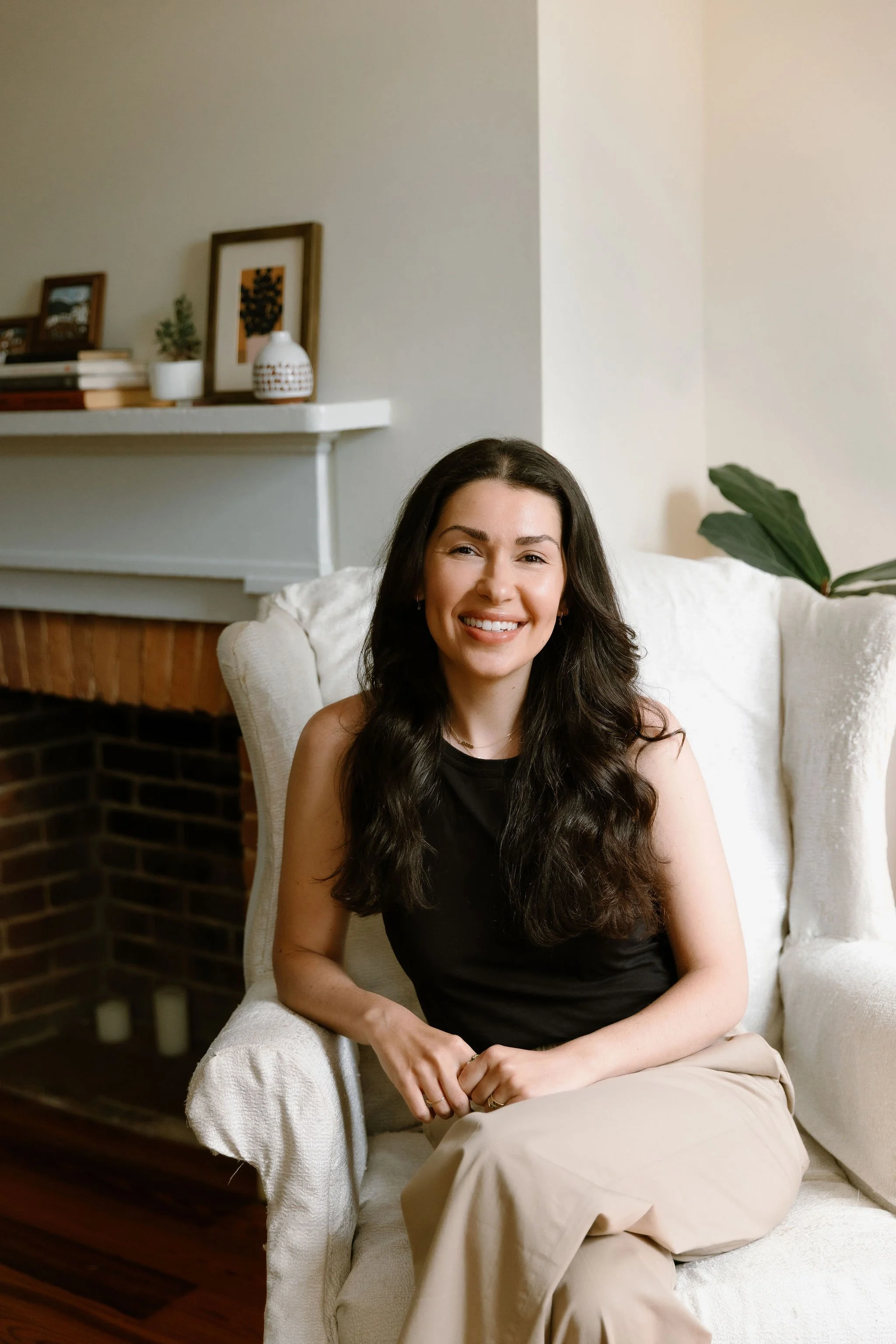 A woman sitting on a white armchair with beige pants and a black top, smiling at the camera. Behind her is a mantel with decorative books, framed artwork, a small potted plant, and a white decorative vase. There is a large green leafy plant to her right.