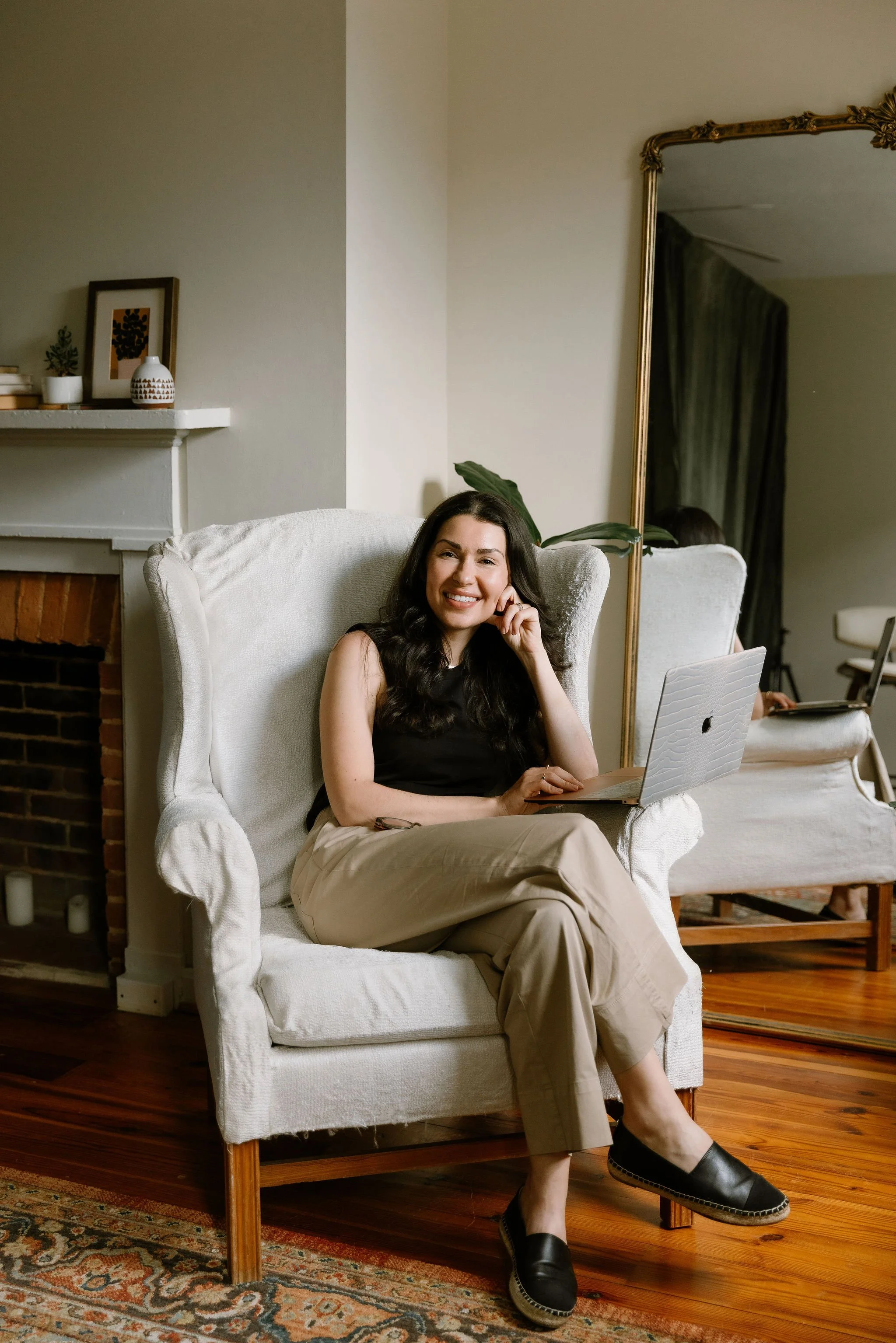 A woman sitting on a white armchair with a laptop on her lap, smiling at the camera in a cozy living room with a fireplace, decorative shelf, a large mirror, and plants.
