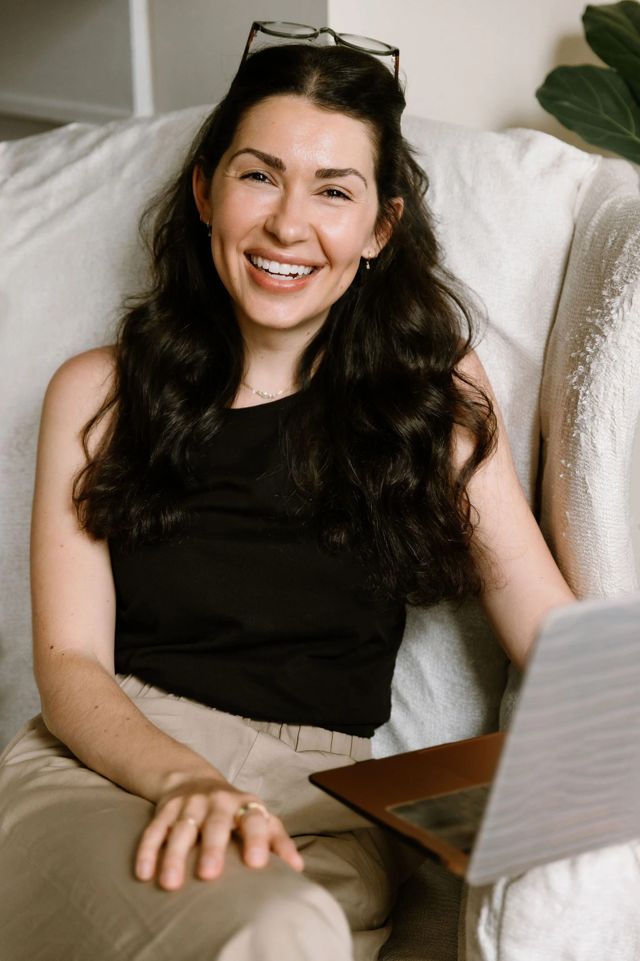 A woman with long dark hair, wearing a black top, sitting on a cream-colored couch, smiling, with glasses resting on her head, and a green plant in the background.