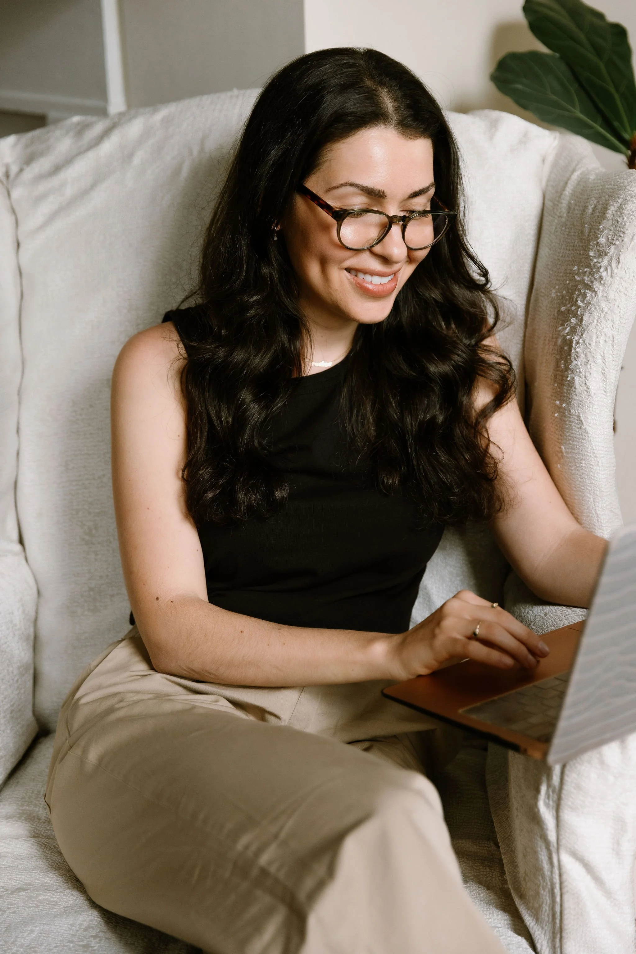 A woman with long dark wavy hair, wearing glasses, a black sleeveless top, and beige pants, sitting on a cream-colored sofa, smiling while using a laptop.