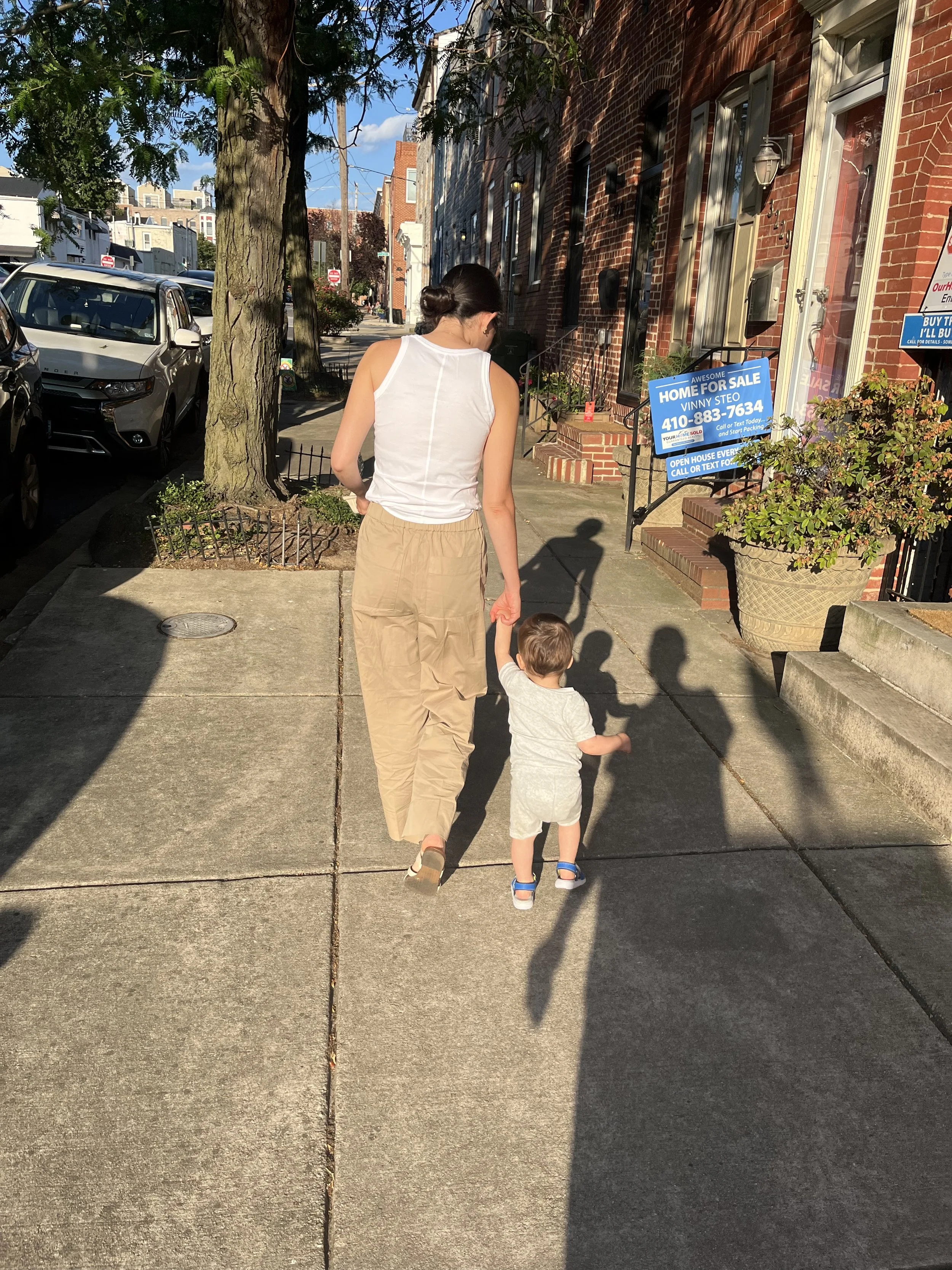 A woman and a young boy walking hand in hand on a city sidewalk in front of brick buildings with signs, trees lining the street, and parked cars on the side.