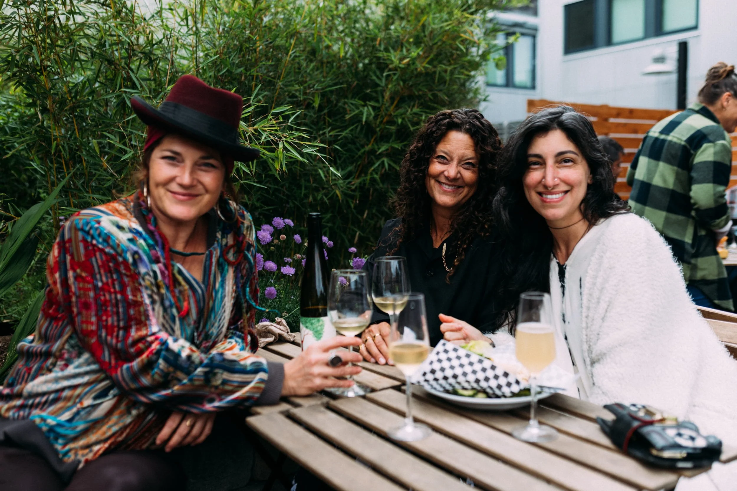 Three people smiling and drinking wine at a wooden bench